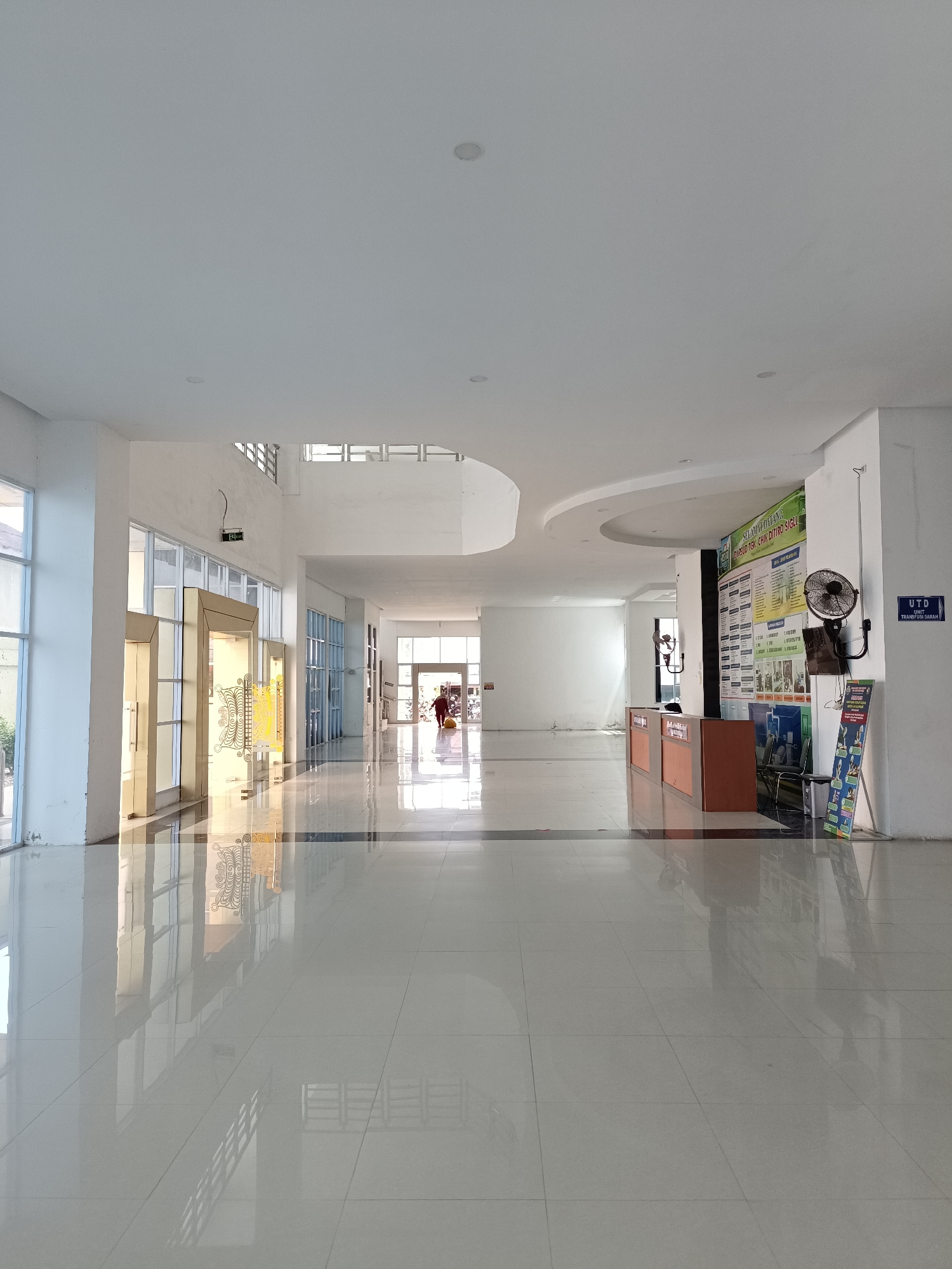 The image depicts a bright, modern, indoor hallway with a high, white ceiling and large windows on either side. The floor is a polished, reflective white tile that shows reflections of the ceiling lights and architectural elements. The hallway extends into the distance, with a slight curve in the ceiling on the right side.
On the right side of the hallway, there is a reception desk with two counters. Behind the desk, a large informational poster is mounted on the wall, featuring text and images. A black oscillating fan is mounted on the wall above the poster. To the right of the poster, a sign reads "UTD UNIT TRANSFUSA DARAH". A small display stand with promotional materials is located to the right of the reception desk.
Further down the hallway on the left, there are large glass doors with ornate golden accents. Through these doors, a glimpse of an outdoor area with greenery is visible.
In the distance, at the end of the hallway, two individuals can be seen. One person, wearing a red shirt, appears to be cleaning or sweeping the floor. The environment suggests a public building, possibly a hospital or administrative center, given the reception desk and the "Transfusa Darah" sign. The overall impression is one of cleanliness and spaciousness. The specific location in Mesjid Reuntoh, Indonesia, is not evident from the visual information alone.