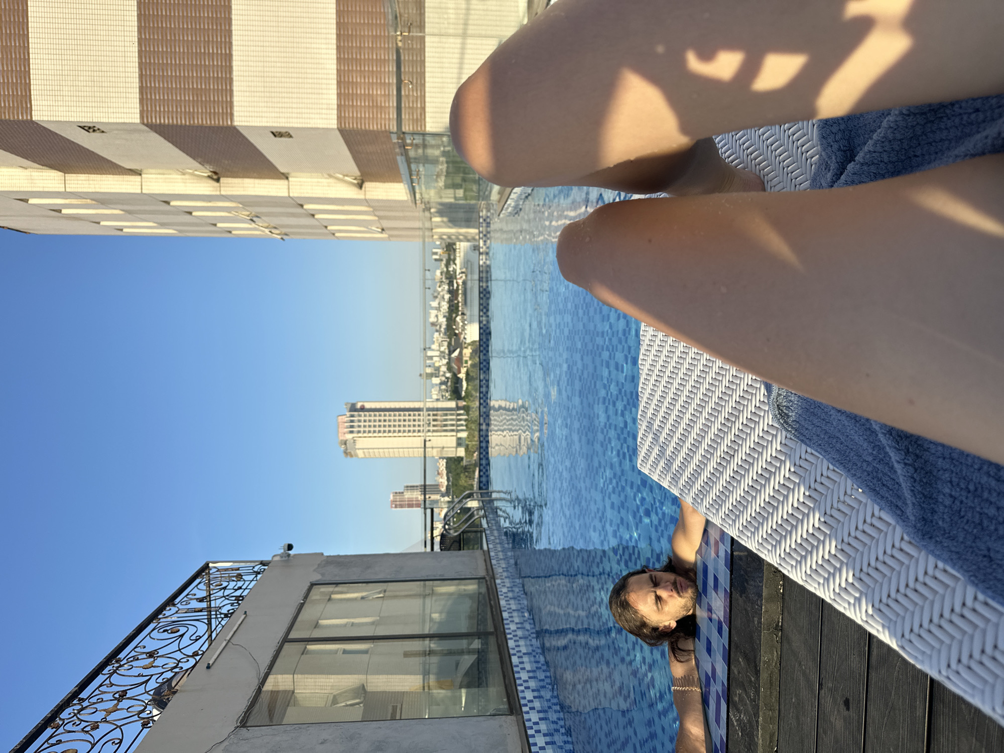 The image shows a person relaxing in a rooftop swimming pool with a city skyline in the background. The person, who appears to be male with long brown hair, is partially submerged in the blue, tiled pool, with their head resting on the edge. They are looking upwards with closed eyes. A portion of their legs is visible, extended out of the water and resting on a white woven lounge chair. A blue towel is draped over the chair.
The setting is a rooftop, indicated by the surrounding buildings and the view of the city. The architecture of the buildings in the background suggests an urban environment, and the presence of high-rise buildings points to a city. The clear blue sky suggests it is daytime, likely sunny with good weather, as indicated by the shadows cast on the lounge chair and the person's legs. The vibrant blue of the pool water and the clear sky contribute to a summery or vacation-like atmosphere.
In the foreground, on the left side of the image, part of the building's facade is visible, featuring a striped pattern in beige and brown tones. Below that, there's a metal railing with intricate scrollwork. Further down on the left, a section of the building's exterior wall and windows can be seen. The pool itself has a checkered blue and white tile border. The overall impression is one of leisure and relaxation in an urban setting. No visible text is present in the image.
