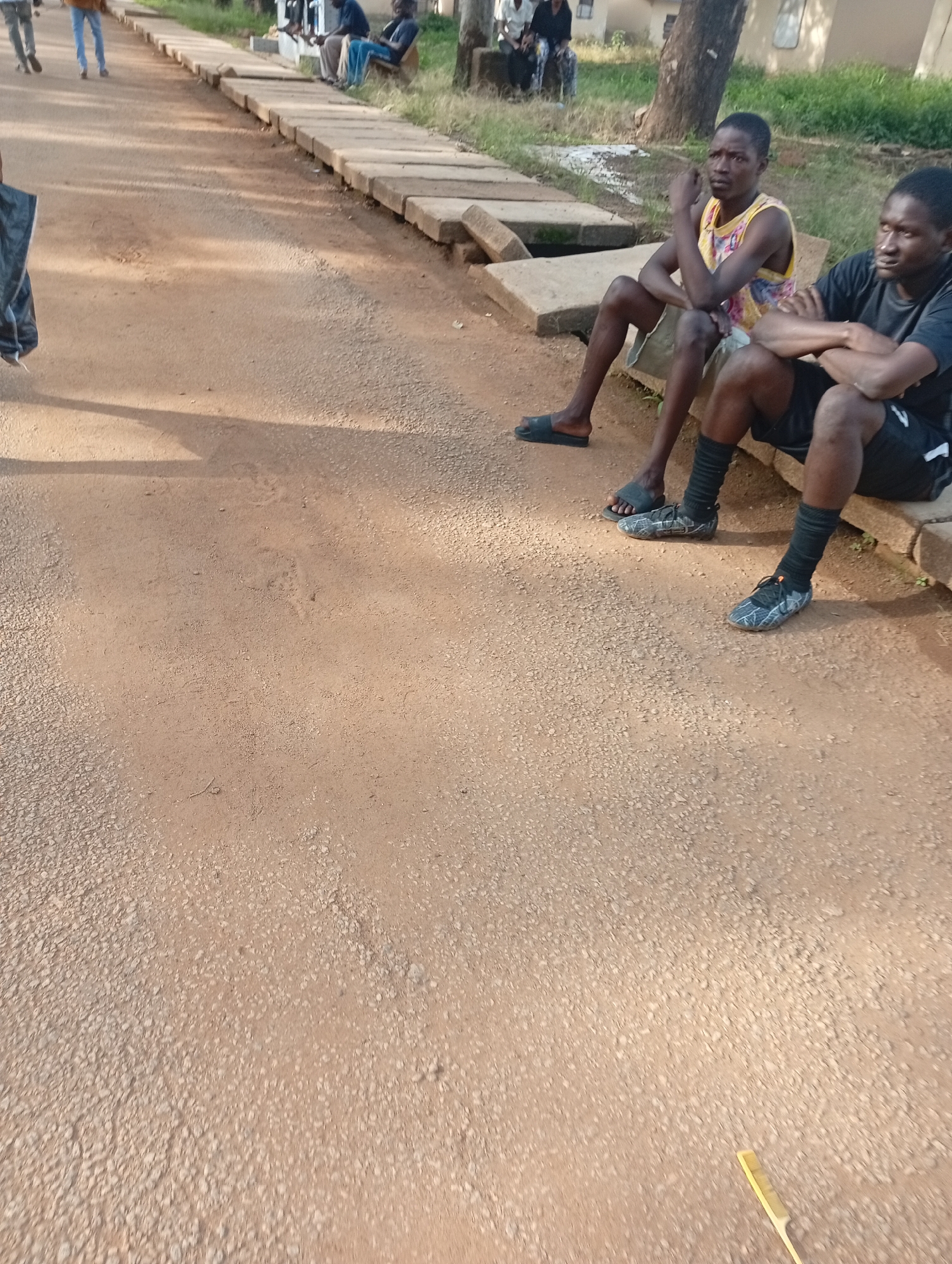 The image shows a dirt road in Rafin Par, Nigeria, with two young men sitting on concrete blocks at the side of the road. The man on the left is wearing a sleeveless, colorful t-shirt and khaki shorts, with black flip-flops. The man on the right is wearing a black t-shirt, black shorts, and black socks with athletic shoes. Both men are looking off into the distance with serious expressions. In the background, other people can be seen walking and sitting, some of whom appear to be children. The scene suggests a casual daytime gathering. The sun is casting shadows on the road, indicating it is likely daytime. The environment appears to be somewhat rural or semi-urban, with trees and buildings visible in the distance. The overall mood is calm and relaxed.