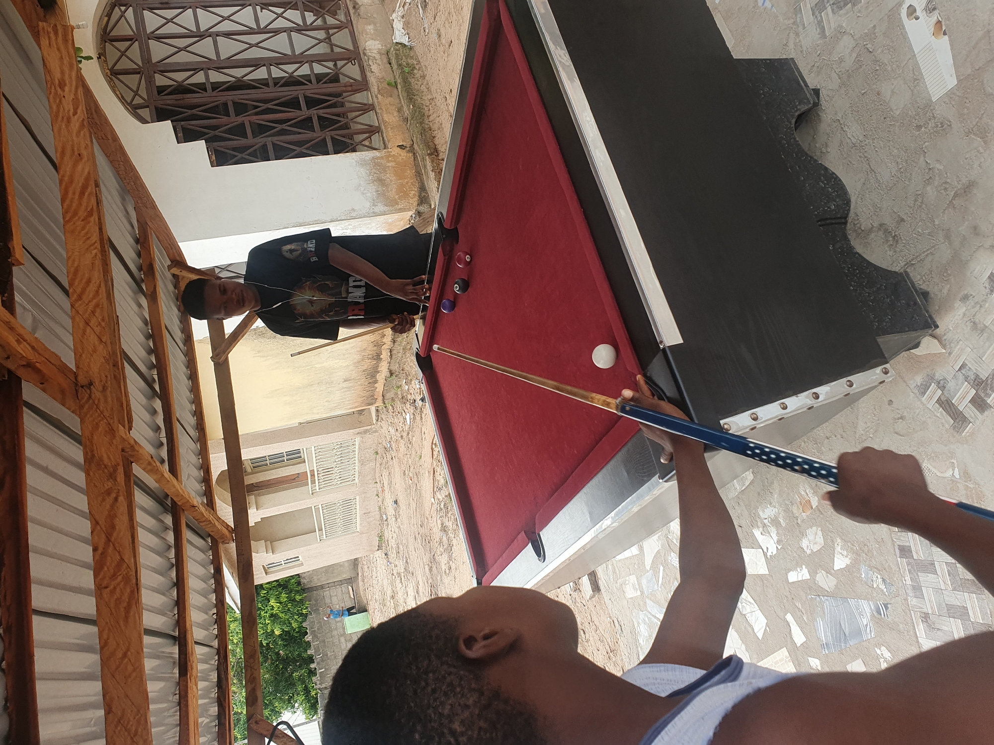 The image depicts two young males playing billiards outdoors in Lokoja, Nigeria. One male, wearing a white tank top, is in the foreground with his back to the camera, holding a billiard cue. The other male, wearing a black t-shirt with a graphic print and headphones, is standing behind the billiard table, also holding a cue. The billiard table is covered with a red cloth and has several colored balls on it, including a white cue ball near the foreground player. The setting appears to be a casual outdoor space with a partially constructed roof supported by wooden beams. In the background, there is a building with a white facade and decorative window grills, and a grey brick wall with some vegetation visible. The ground is a mix of sand, concrete, and scattered tiles.