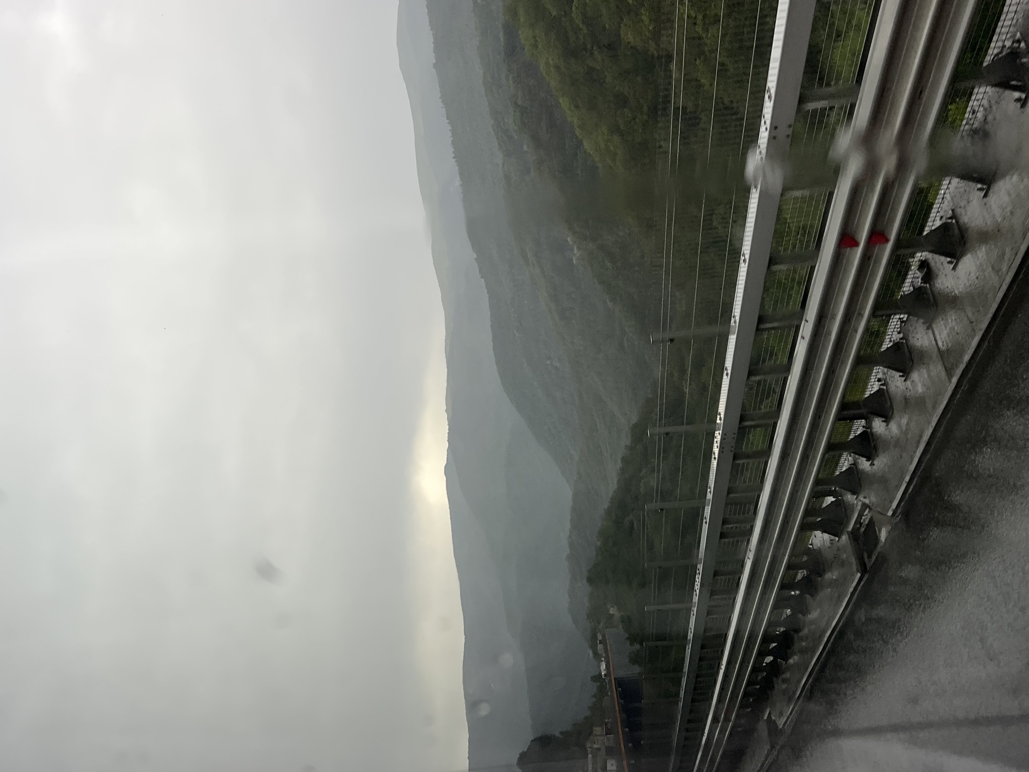 The image depicts a scenic mountain landscape in Gorno Kamartsi, Bulgaria, viewed from a highway overpass. The sky is overcast, suggesting it is daytime with cloudy or rainy weather, as evidenced by water droplets on the visible guardrail. The mountains, covered in lush green trees, recede into the distance, shrouded in a misty haze. The guardrail, made of metal and concrete, runs along the right side of the frame, indicating the perspective of someone traveling on a road. Two small, triangular red markers are visible on the guardrail, potentially for visibility or marking. The overall mood is serene and somewhat somber due to the weather and the vastness of the landscape. No people are visible in the image.