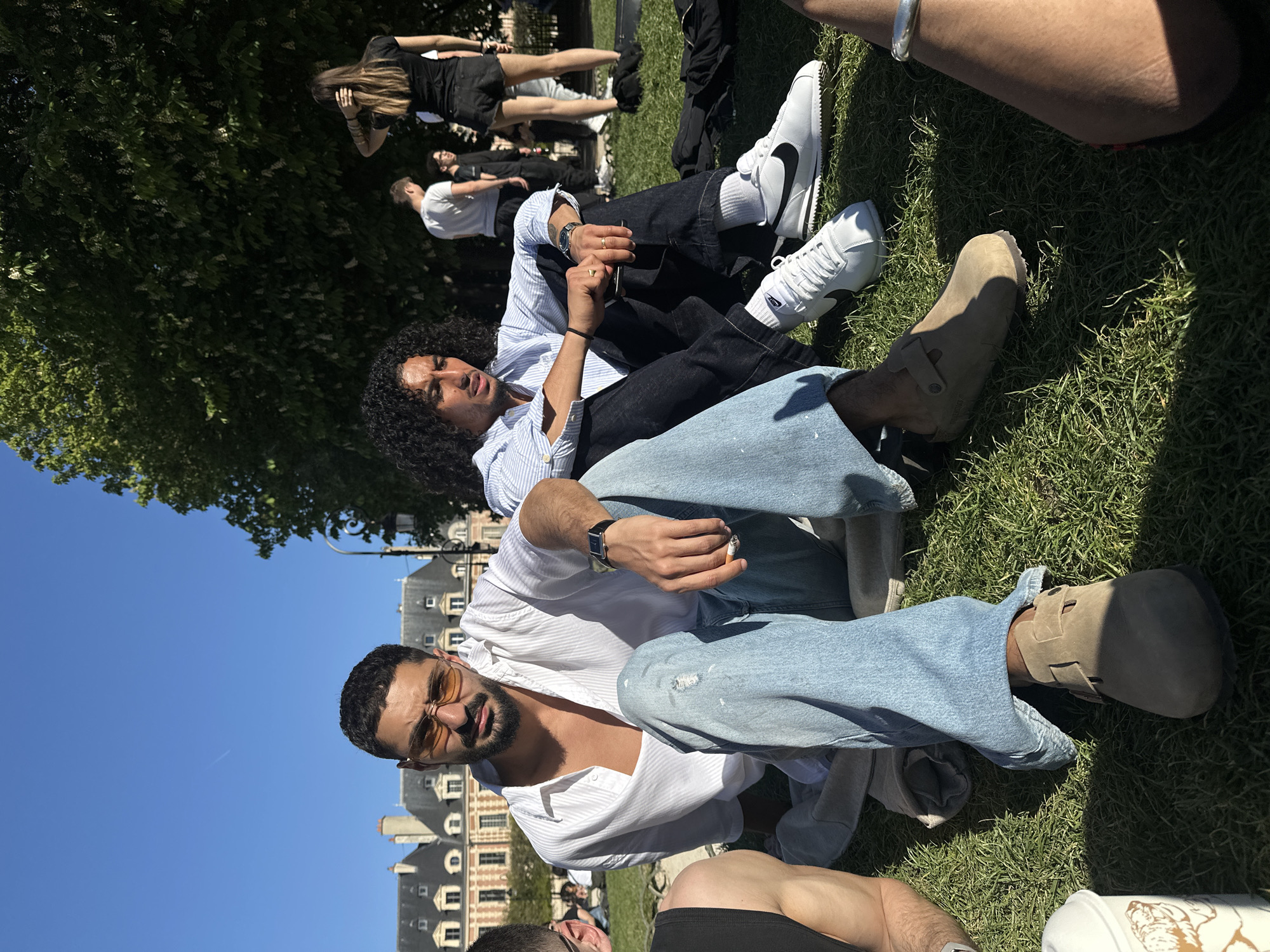 The image shows two men sitting on grass, seemingly in a park in Paris, France. One man wears a white, unbuttoned shirt, light blue jeans, brown sandals, and sunglasses, while the other wears a light blue striped shirt, dark jeans, and white sneakers. The man in the white shirt sits in front. Both men are wearing watches. The background reveals buildings typical of Parisian architecture and other people relaxing on the grass. The sky is clear and blue.