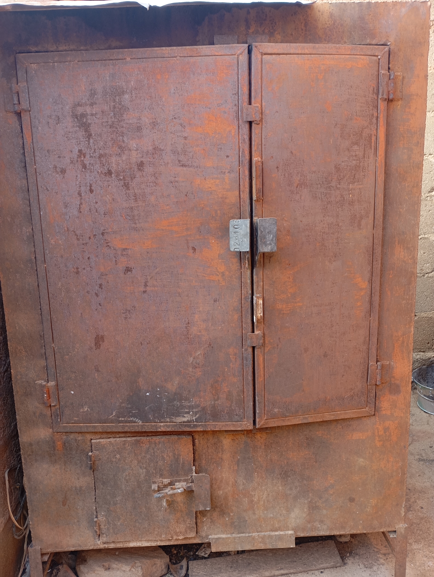 An outdoor, medium shot captures a heavily rusted, large metal cabinet or oven with double doors. The cabinet is a dull reddish-brown due to extensive rust. The doors are framed by metal and have visible hinges on the left and right sides. A metal latch with what appears to be a small plaque or tag reading "02-103" is visible on the seam between the two doors, securing them closed. Below the double doors, there is a smaller, square door with a latch. The cabinet sits on what appears to be a concrete or dirt surface, with some rocks and debris visible around its base. The background shows a textured, light-colored wall, possibly brick or concrete, and a glimpse of a metal bucket on the right side of the frame. The overall scene suggests an industrial or utilitarian setting, possibly in a yard or workshop.