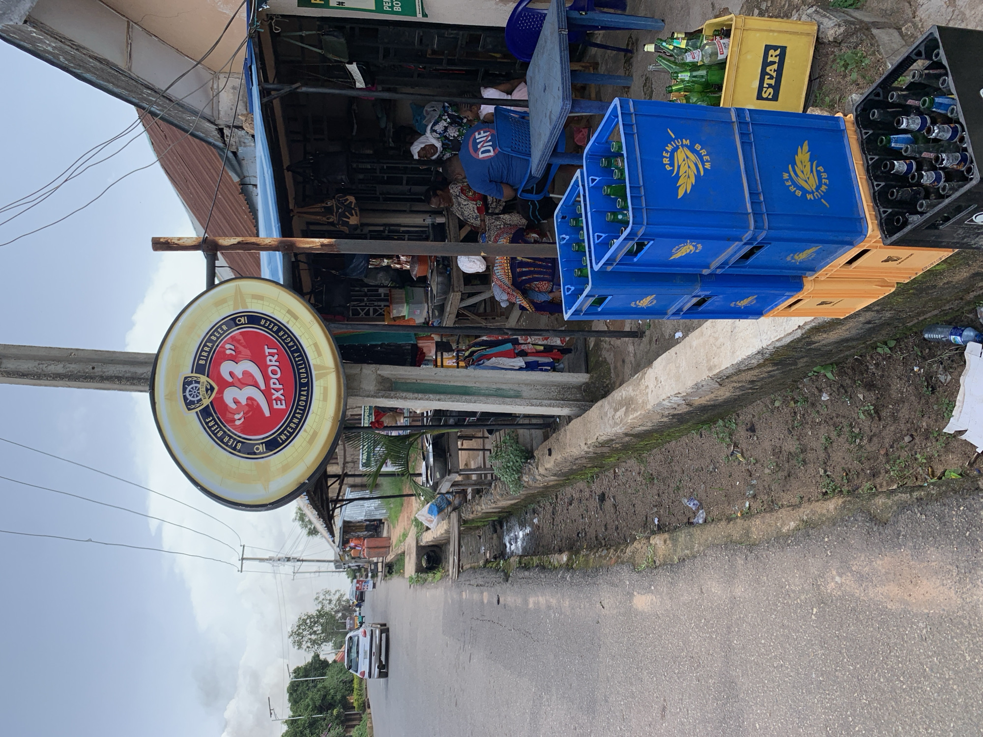 This media file depicts an outdoor scene in what appears to be a market or roadside stall area, likely in Jos, Nigeria. The main subjects are several people, presumably customers and vendors, gathered around a stall. The setting is a street with a paved road, a raised concrete curb, and various structures that suggest a commercial area.
Prominently displayed is a large circular sign advertising "33" Export International Quality Lager Beer. To the right of the stall, several blue plastic crates filled with green beer bottles are stacked, along with a yellow crate labeled "STAR". Another black crate, also filled with bottles, is partially visible. The scene also includes a white car parked on the road, and other vehicles in the distance.
The activity seems to be that of a typical day at a roadside business, with people interacting and goods being displayed for sale. The lighting suggests it is daytime, and the sky appears overcast or cloudy, indicating possibly humid or uncertain weather conditions. The presence of the beer crates and the signage points towards a beverage sales point, possibly a small bar or shop. Some of the people are seated on chairs, engaged in conversation. The overall atmosphere appears casual and functional, indicative of a busy local commercial environment. Visible text includes brand names like "33" Export, "STAR", and "PREMIUM BREW", as well as "DNF" on a person's shirt. The scene captures a slice of everyday life in a Nigerian commercial street setting.