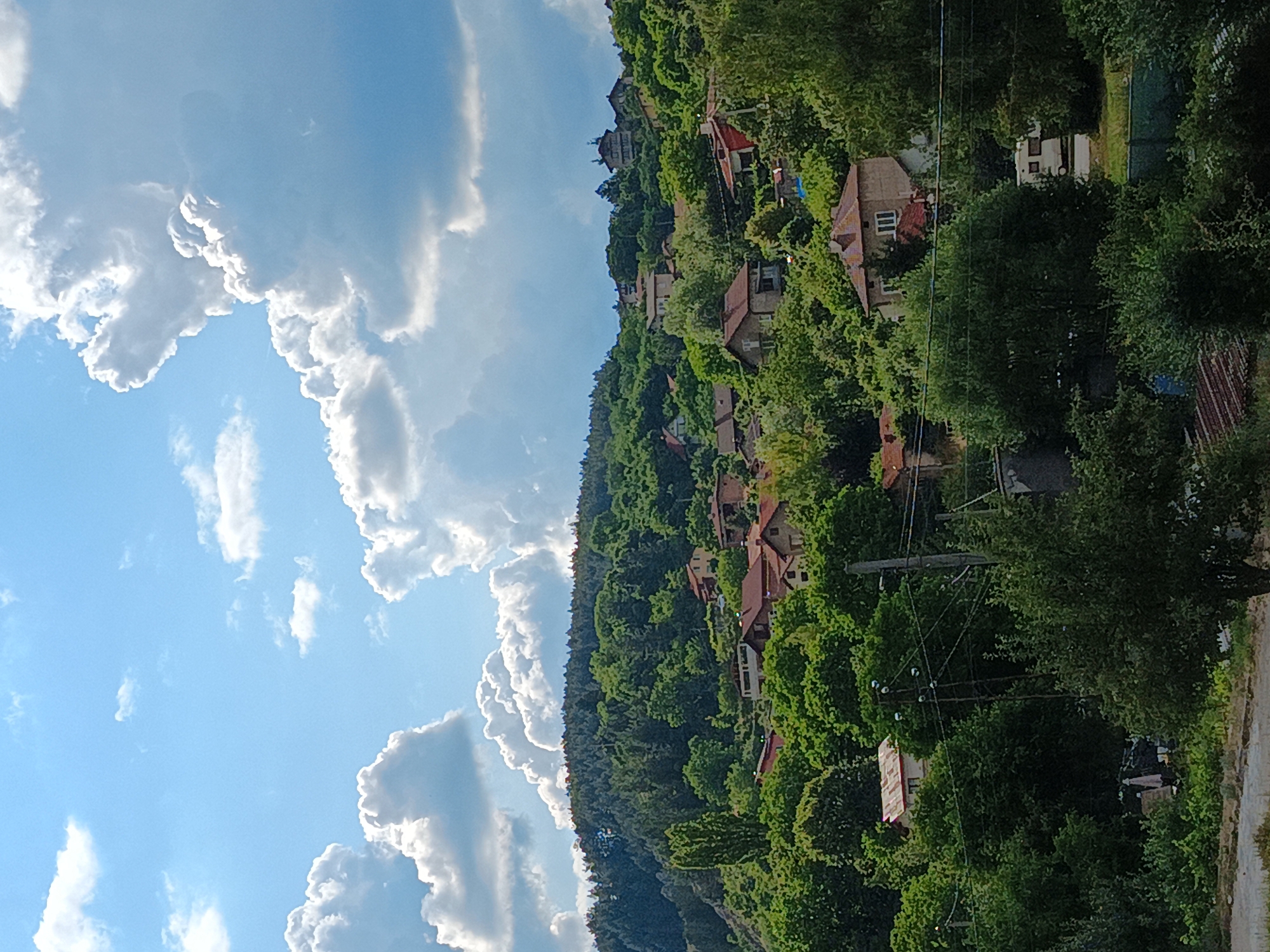 This media file depicts a hillside village in Kralev Dol, Bulgaria. The scene is set on a bright, sunny day with scattered white clouds against a clear blue sky. The hillside is densely covered with lush green trees, interspersed with a scattering of houses with red-tiled roofs. The houses appear to be traditional European village homes, some with visible windows and chimneys.
A prominent feature of the scene is a series of utility poles with wires running across the hillside, indicating the presence of infrastructure within the village. The houses are nestled amongst the trees, suggesting a close integration of the settlement with its natural surroundings. In the foreground, a rough, unpaved road or path is visible, hinting at the access routes to and within the village.
The overall impression is one of a peaceful, rural setting, bathed in the warm light of the daytime sun. There are no people or specific activities visible in the frame, focusing the attention on the landscape and the architectural elements of the village. The location cues, such as the architecture and the name "Kralev Dol, Bulgaria," firmly place the scene in Eastern Europe. The weather appears pleasant, with ample sunlight and fluffy clouds, suggesting a good day for outdoor activities, though none are captured here. The image conveys a sense of tranquility and the rustic charm of a village built into a hillside.