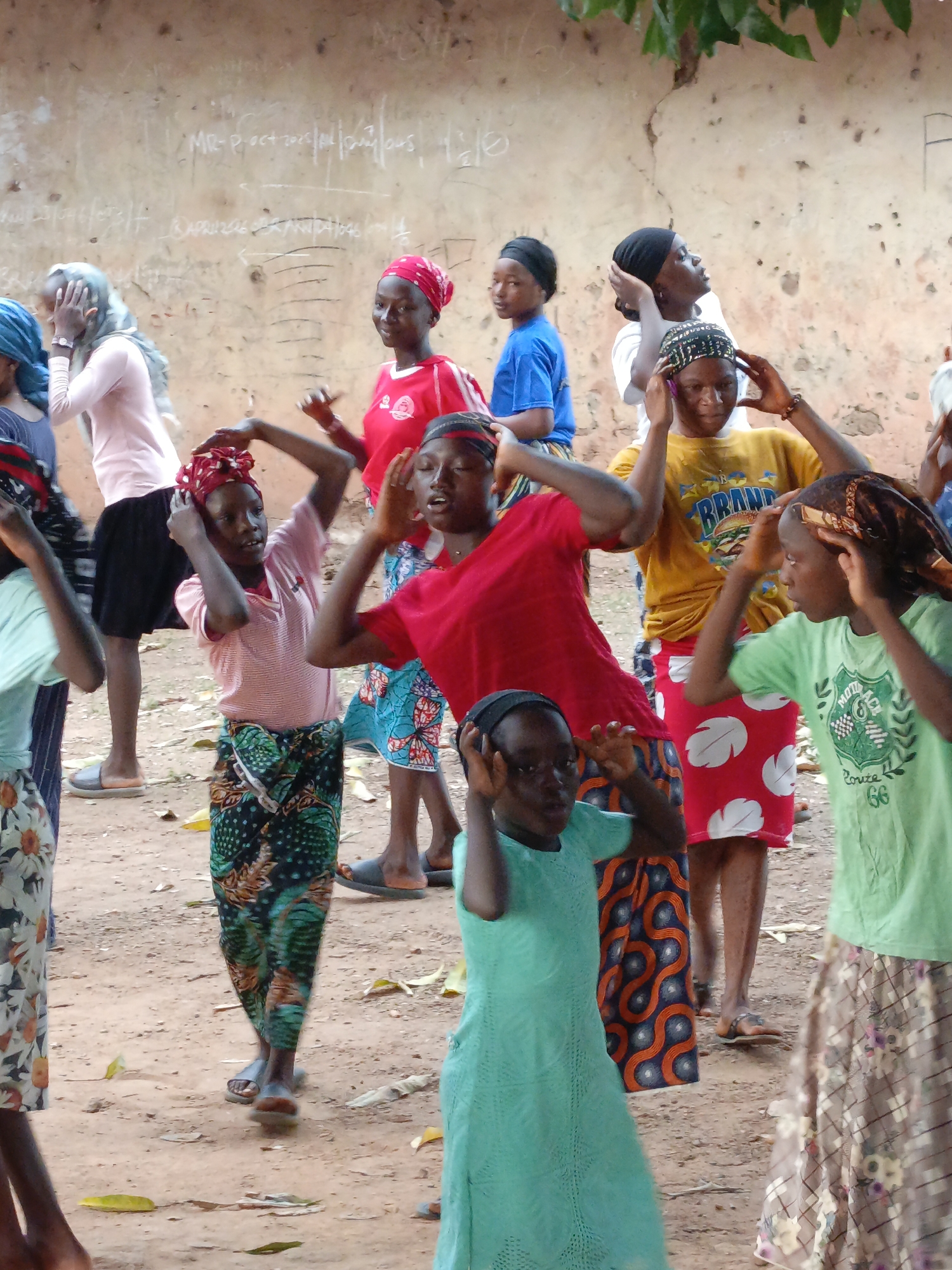 A group of approximately 15 individuals, predominantly girls and young women, stand on a dirt ground in what appears to be an outdoor courtyard in the city of Akwanga, Nigeria. They are in front of a pale building with text written in chalk on the wall. The people are wearing casual clothing in bright colors and some have head coverings or headbands. Most are holding their hands on their heads.