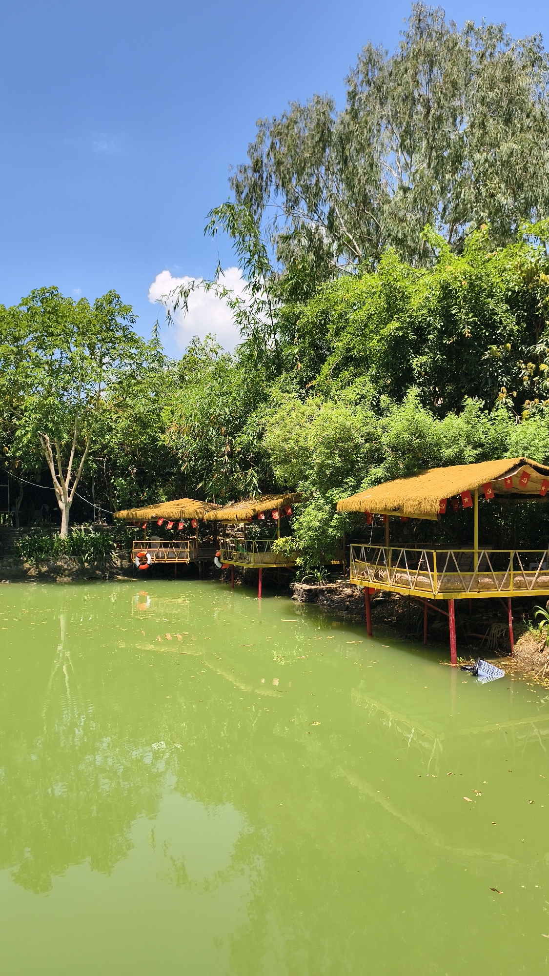 The image depicts an outdoor dining area by a green body of water, likely a lake or pond, surrounded by lush vegetation and trees under a clear blue sky with a few scattered clouds. There are several elevated, thatched-roof huts with yellow railings situated along the water's edge, supported by red poles. The huts appear to be designed for dining or relaxation, with seating visible inside some of them. Red lanterns and flags are hung from the huts. The water reflects the trees and structures, indicating calm conditions. The general environment suggests a serene, natural setting, possibly a restaurant or resort. The location is identified as Nam Nha Trang Ward, Vietnam.