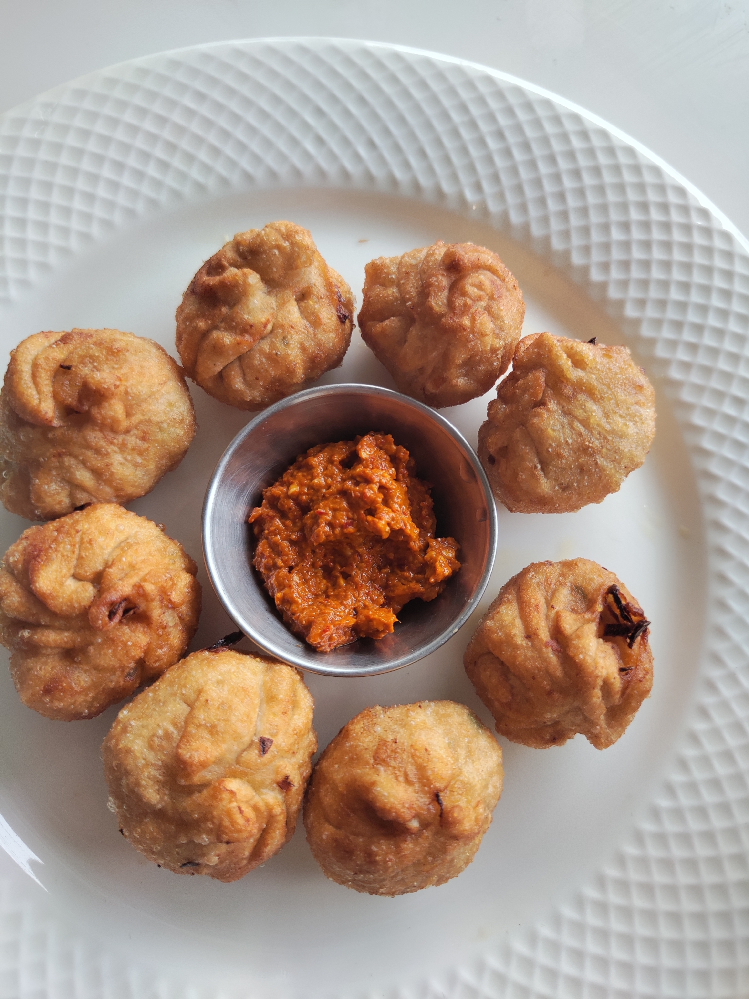 A top-down, close-up shot shows a white textured plate containing eight fried dumplings arranged in a circle around a small metal bowl filled with a red-orange paste. The dumplings are golden brown and have a wrinkled appearance, suggesting they are possibly momos. The paste in the bowl appears to be a chili-based sauce or chutney. The plate rests on a light-colored surface. The image was captured in Dharamkot, India.
