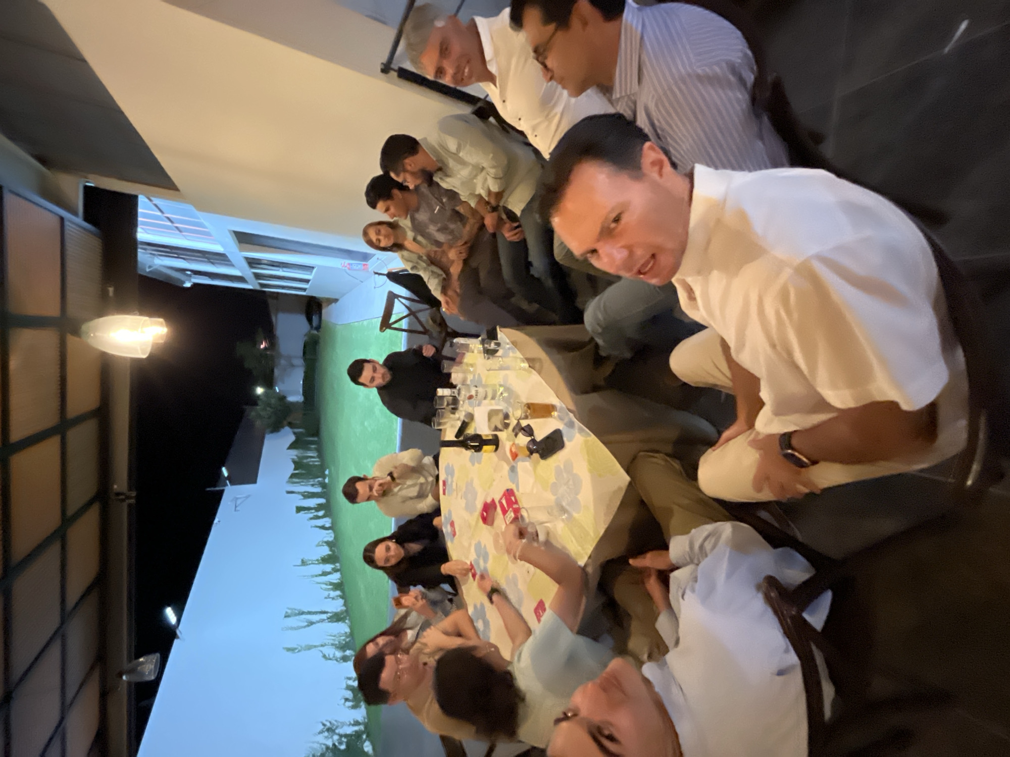 A group of approximately 15 people are gathered around a table, seemingly enjoying a meal or social gathering. The table is covered with a floral patterned tablecloth, with glasses, bottles, and other dining items scattered across it. Several individuals are engaged in conversation or interacting with each other. The environment appears to be an outdoor patio or courtyard, with a building visible in the background. The location is in San Bernardino Tlaxcalancingo, Mexico.