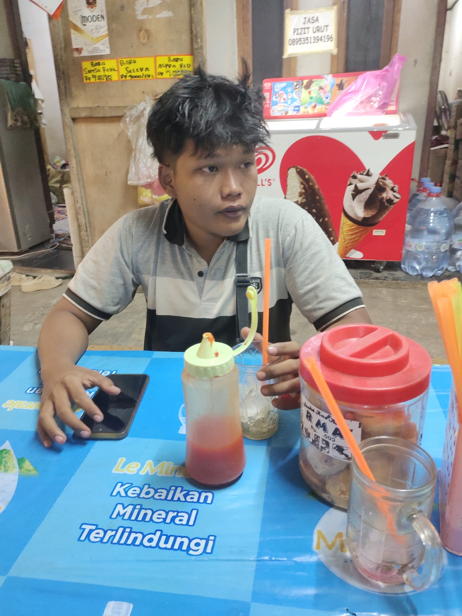 The image depicts a young man sitting at a table in what appears to be a small eatery or food stall in Lengkong Kulon, Indonesia. The man is wearing a gray and black polo shirt and has dark, messy hair. He is holding a smartphone in his left hand, seemingly browsing or using it.
In front of him, on a blue tablecloth, are several objects. To his left, there is a clear bottle with red liquid, likely a sauce or condiment, with a yellow cap. Next to that is a clear glass with a straw, containing ice and a light-colored liquid. To his right, a large clear jar with a red lid contains some round, light-brown items, possibly biscuits or snacks. Another glass with an orange straw is positioned next to this jar. An orange straw is also visible in the jar. In the background, an upright freezer with images of ice cream is visible, along with some bottled water. A sign with Indonesian text, including a phone number, is also present in the background, suggesting services are offered. Other posters with text are on the wall behind the man. The overall impression is a casual, everyday scene in a local Indonesian establishment.