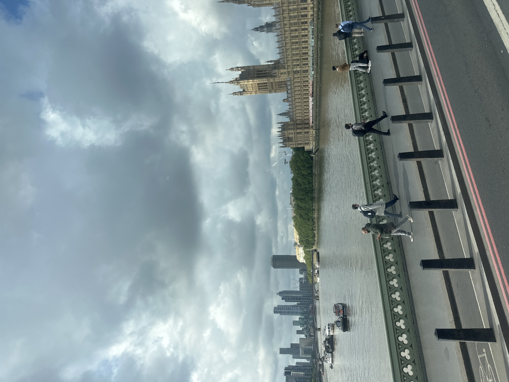 The image depicts a view over the River Thames in London, United Kingdom, looking towards the Palace of Westminster (Houses of Parliament). The perspective appears to be taken from a bridge, potentially Westminster Bridge or Lambeth Bridge, with pedestrians visible on the sidewalk. The river is filled with boats. The sky is overcast with grey clouds. The city skyline is visible in the distance, comprising modern buildings.