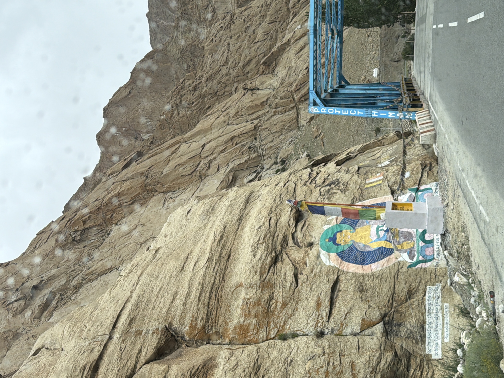 A large, painted mural of a seated Buddha is depicted on a rocky cliff face in Likche, India. The mural features the Buddha in a golden robe, seated on a blue lotus. To the right of the mural are prayer flags and a small white stupa. Below the mural, there is text written in white on the rock face. A blue bridge with the words "PROJECT HIMALAYAS" written vertically on its support beams spans a road in the background. The sky is overcast, and there are water droplets on the camera lens. The environment appears to be mountainous, with rugged terrain and sparse vegetation.