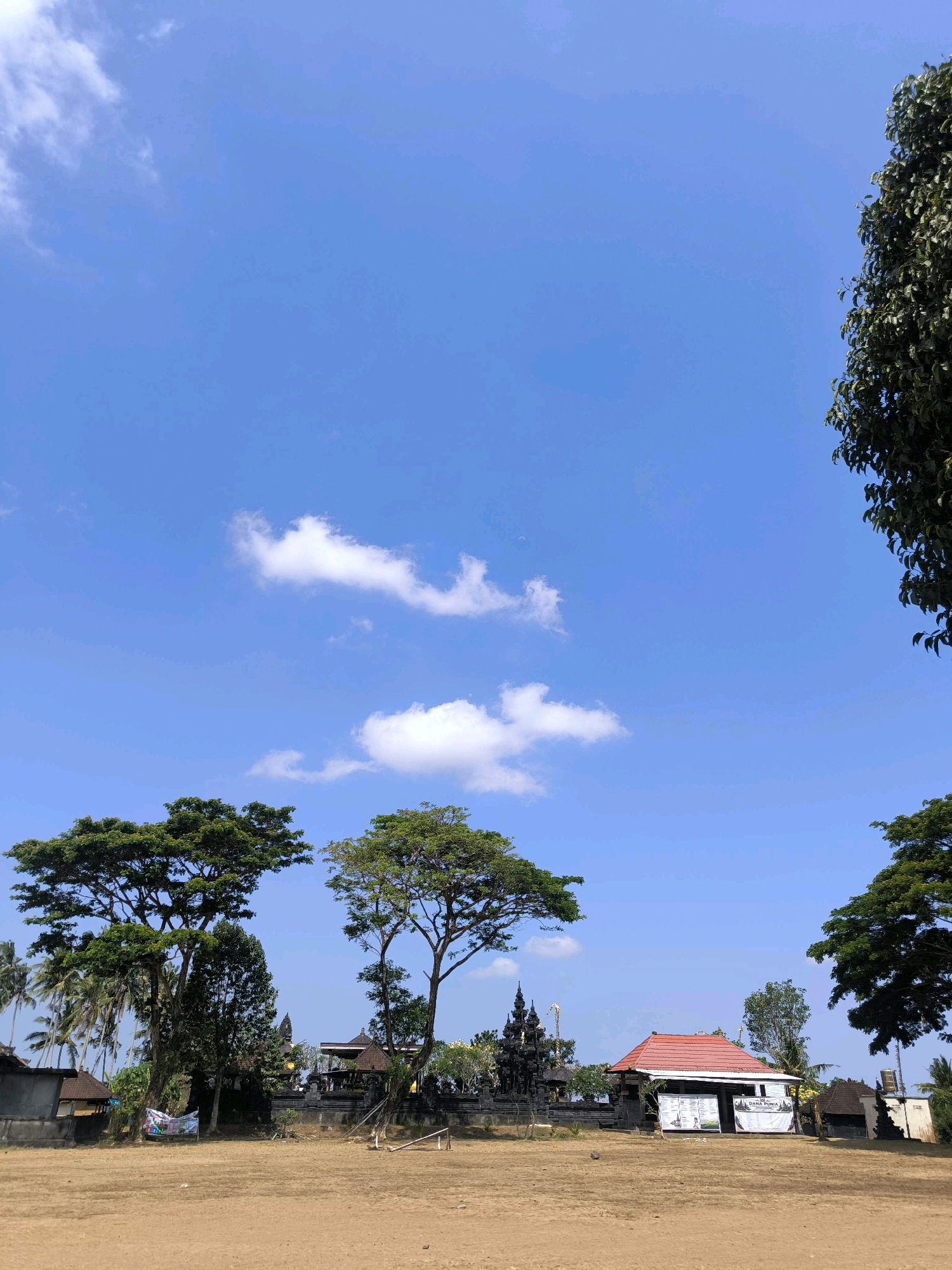 The image captures a bright, sunny day in Tabanan, Indonesia, with a clear blue sky dotted with a few white clouds. In the foreground, a dry, sandy field stretches out, with some sparse vegetation and what appears to be a small, weathered wooden structure or marker in the center. To the left and right, large trees with lush green foliage frame the scene.
In the mid-ground, a traditional Balinese temple complex is visible. It features intricate dark stone architecture, including tiered structures with pointed roofs and decorative carvings. Adjacent to the temple, a building with a prominent red tiled roof stands out. Banners and signs are displayed on and around this building, suggesting it might be an information center or part of a cultural attraction. Palm trees are scattered throughout the background, indicating a tropical setting. The overall atmosphere is serene and suggests a location of cultural or historical significance.