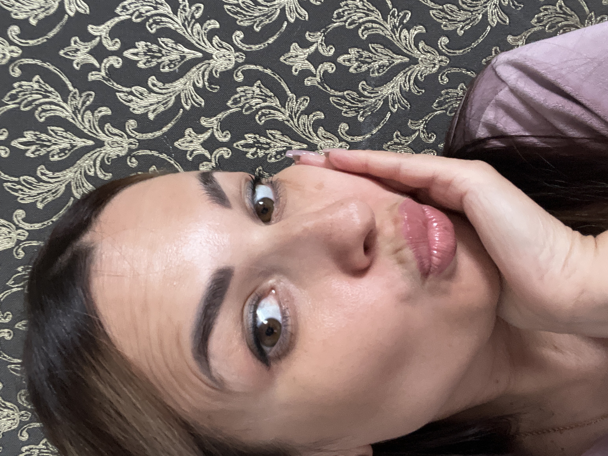 A close-up selfie shows a woman with dark hair and brown eyes pouting her lips. She is resting her right hand on her cheek, with her index finger extended upwards, touching her nose. Her left eye is looking towards the camera, while her right eye is slightly averted. The woman is wearing a light purple top. The background is a dark patterned wallpaper with a gold damask design. The scene is indoors.
