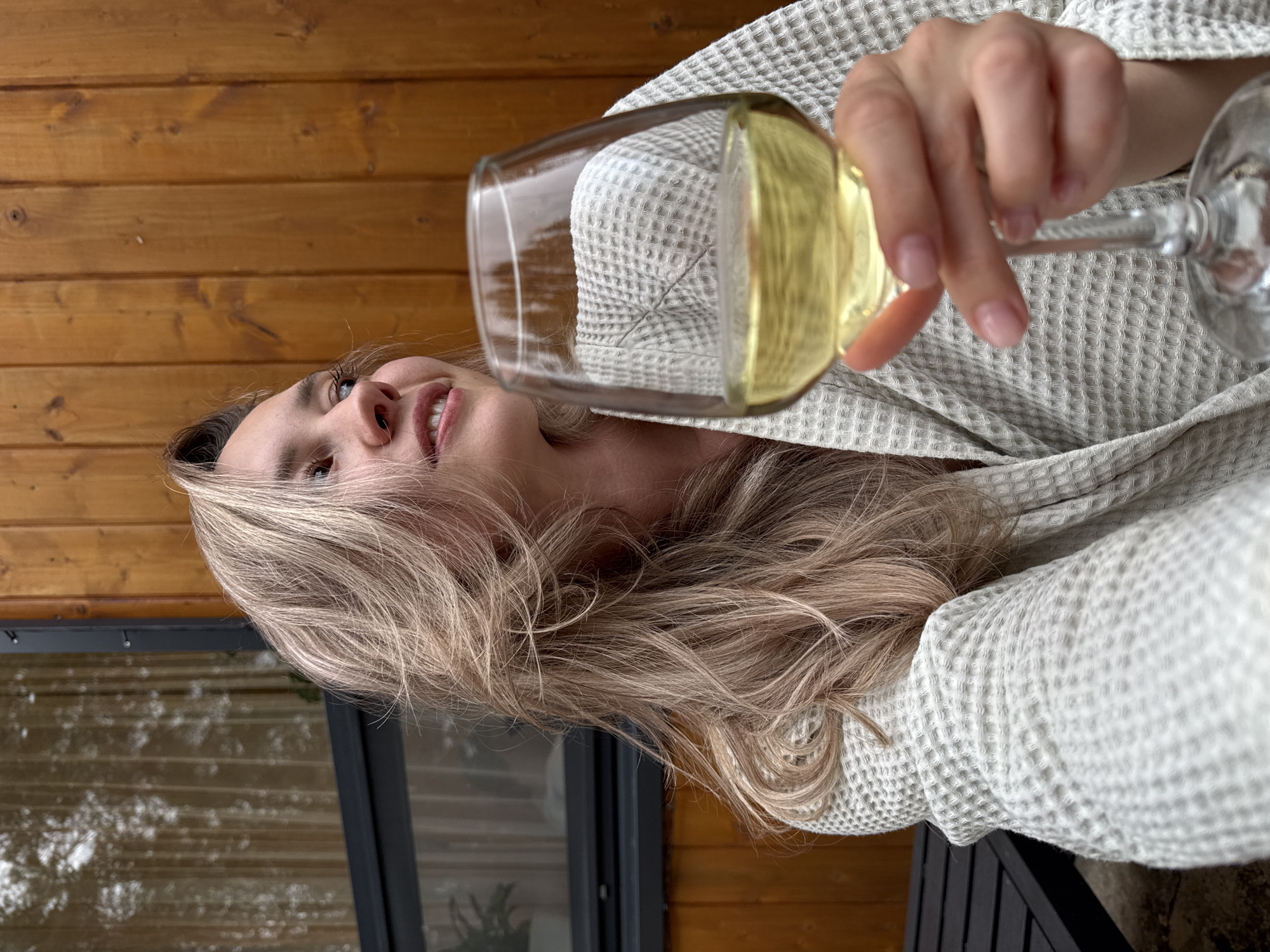 A blonde woman in a white waffle-patterned robe holds a wine glass with yellow liquid. She is outdoors with a wooden wall behind her and a window below. This location is unknown in the city of Chaykovsky, Russia.