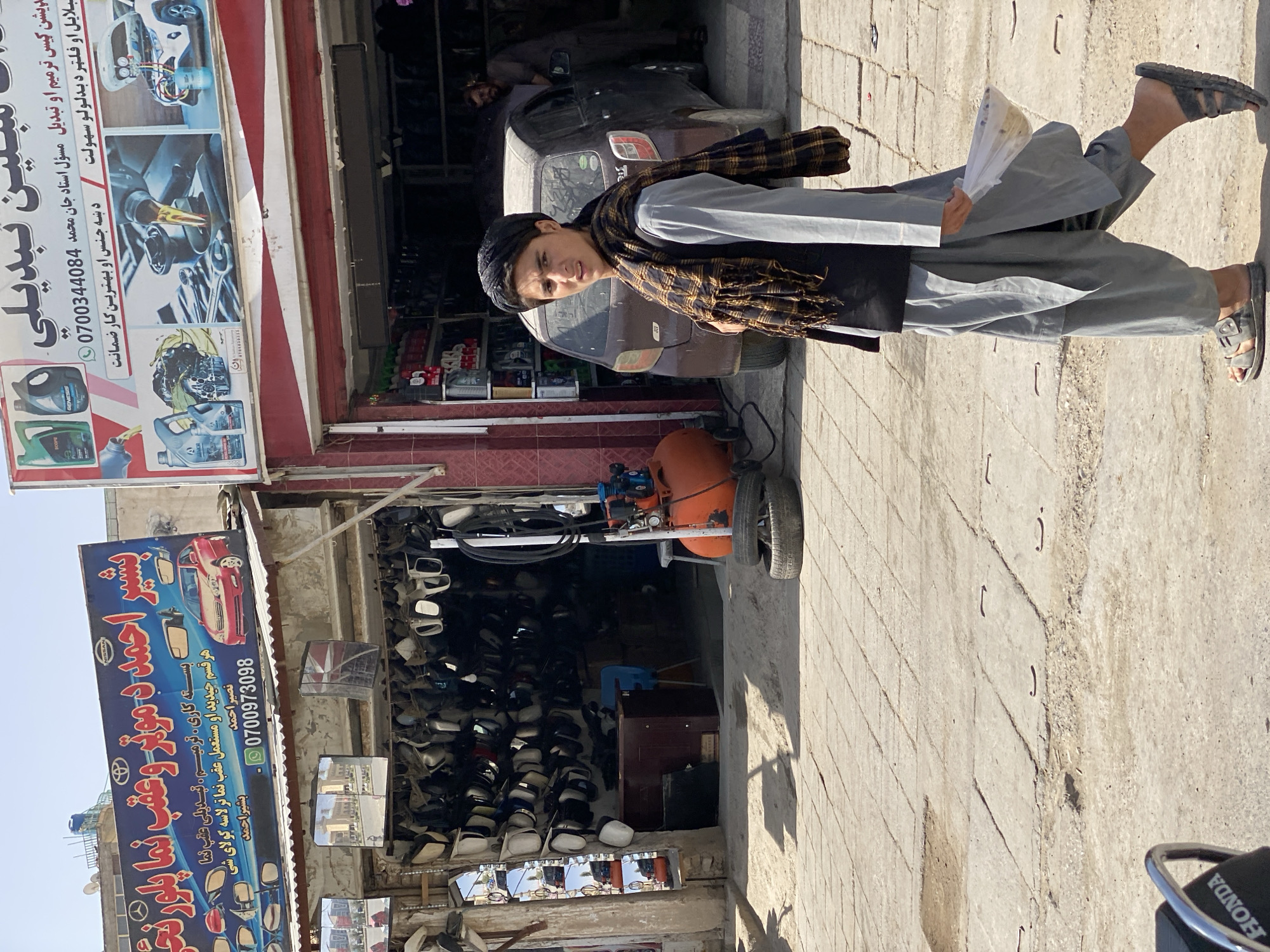 A young male wearing a black turban, a black vest over a grey tunic, and grey pants is walking in front of a shop in Kandahar, Afghanistan. He is also wearing a plaid scarf and sandals, and holding a white plastic bag. Behind him, a brown car is parked in front of another shop that displays various car mirrors. To the left of the main shop, a sign in Arabic advertises car parts. To the right, a sign in Arabic advertises oil change and other car services. An orange air compressor and two tires are visible in front of the shop. The ground is paved with concrete slabs.