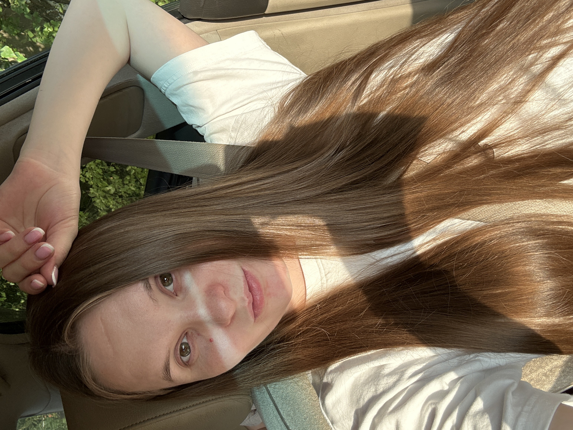 A woman with long, brown hair and green eyes is sitting in the passenger seat of a car, possibly in Noviy, Russia. She is wearing a white t-shirt and has her right arm raised, with her hand resting on her head. Sunlight is casting a diagonal shadow across her face and upper body. The car's interior, including a seatbelt and part of the seat, is visible in the background. Outside the car, foliage can be seen, suggesting a natural, possibly urban fringe, environment.