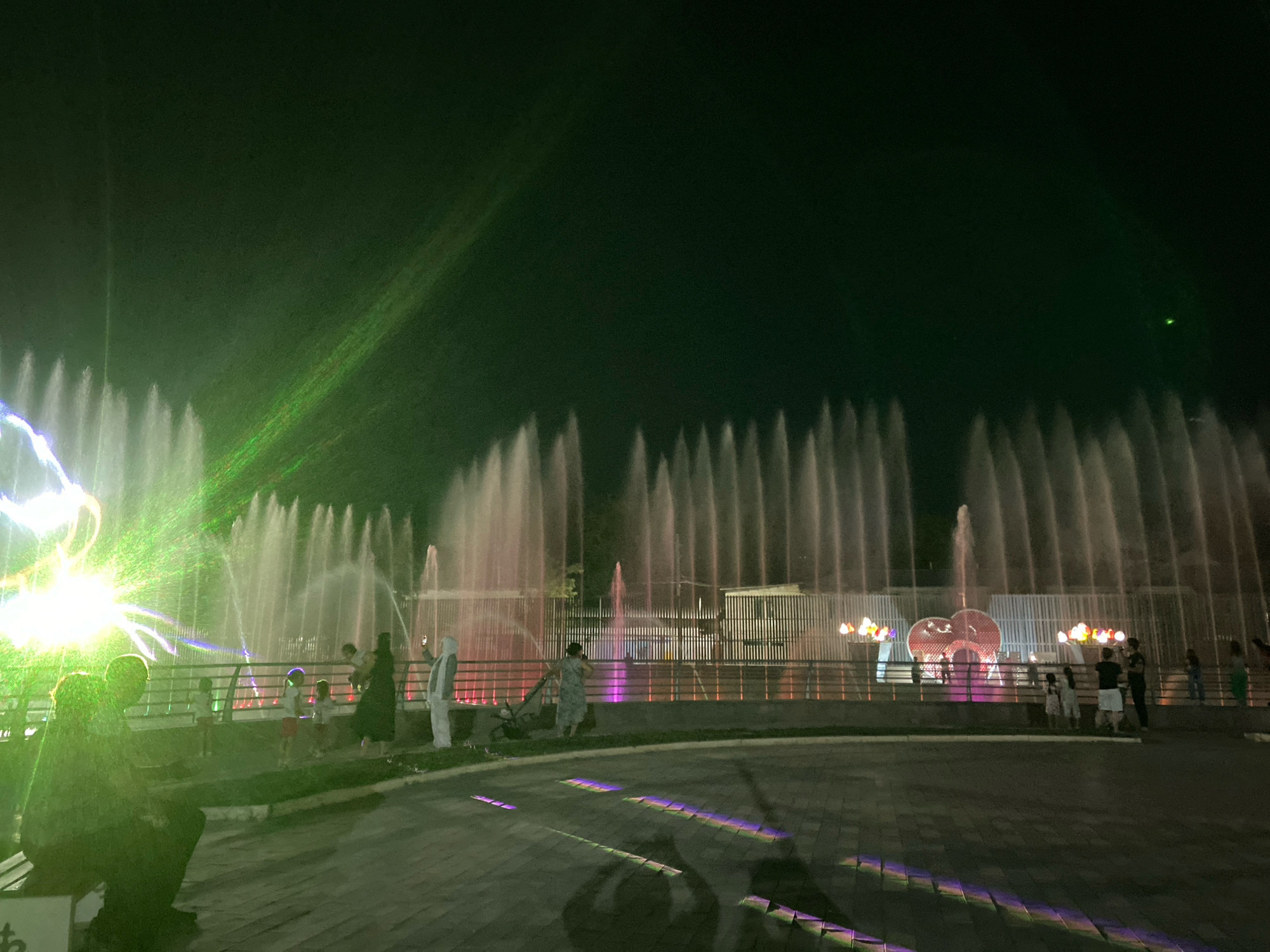 The image depicts a night view of a synchronized water fountain show in Shymkent, Kazakhstan, likely at the "Orda" Fountain. The scene is illuminated by colorful lights and lasers projected onto the water jets, creating a vibrant spectacle. In the foreground, silhouettes of people are visible, some seated on a bench and others standing, observing the show. Further back, a group of people, including children, are gathered near a railing, watching the fountains. The water jets are tall and graceful, forming various patterns, while the background features illuminated structures and more fountains. The overall atmosphere is festive and engaging.