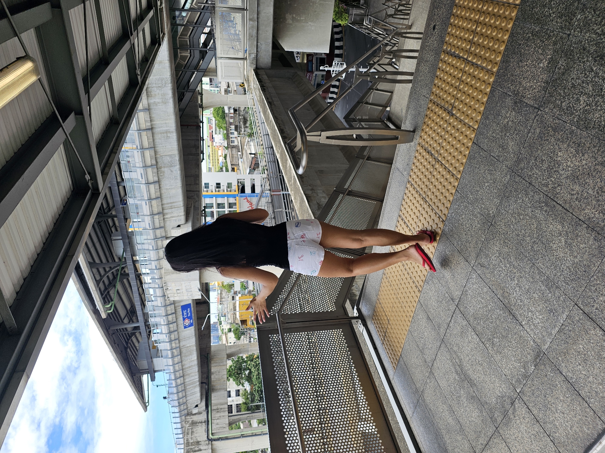 A woman with long dark hair, wearing a black tank top and white patterned shorts, stands facing away from the camera on a covered outdoor walkway in Bangkok, Thailand. She is wearing red flip-flops. The walkway is paved with grey granite tiles and features a tactile paving strip for the visually impaired. A metal railing with a perforated design separates the walkway from a lower level. In the background, an elevated train line runs overhead, with a concrete support structure and tracks visible. Buildings and trees can be seen in the distance. The sky is cloudy. The scene appears to be a public transportation station.