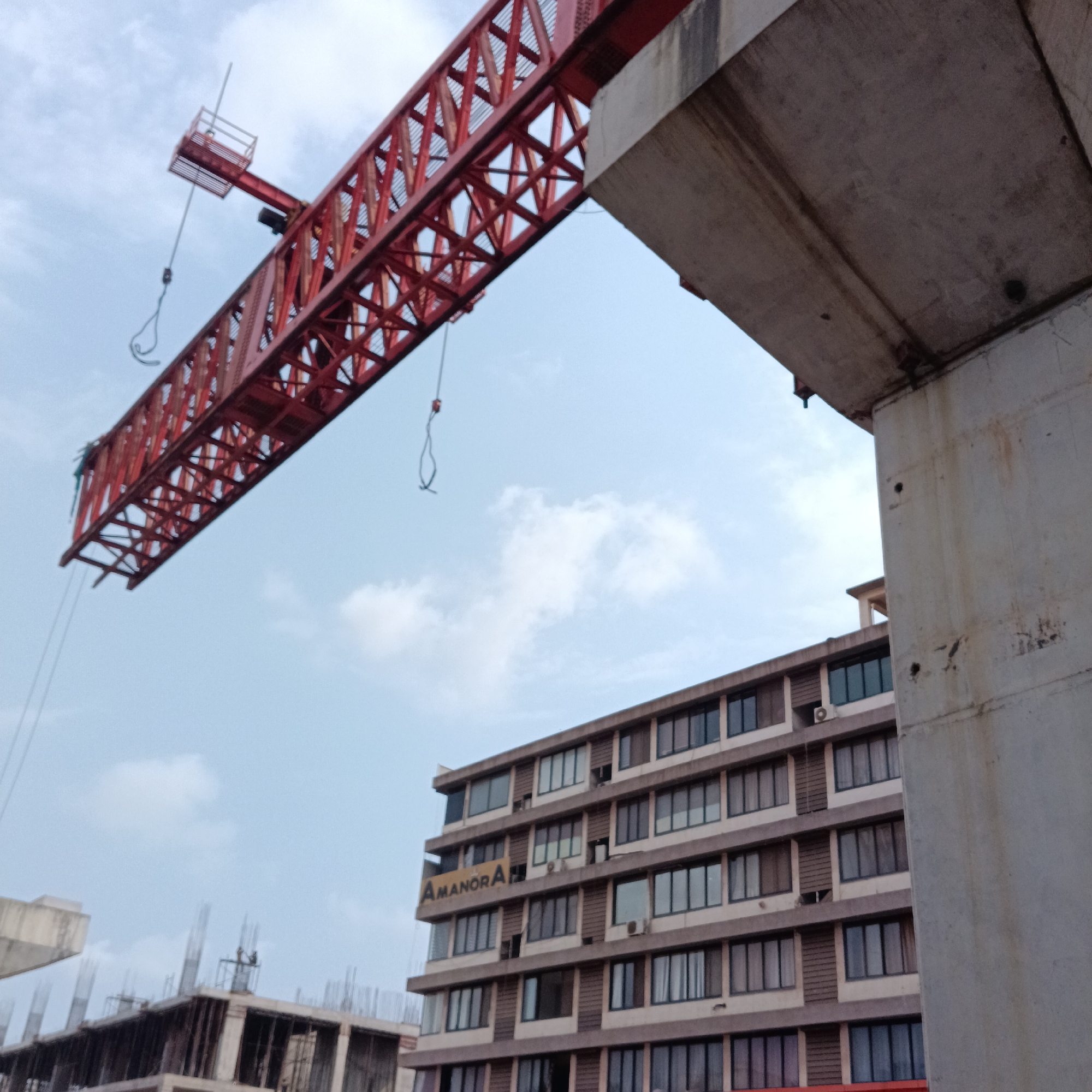The image shows a construction site in Sangolda, India. A red crane arm extends over a partly constructed building, adjacent to an established building labeled "Amanora". A large concrete support structure is visible in the foreground. The sky is overcast. No people are clearly visible.