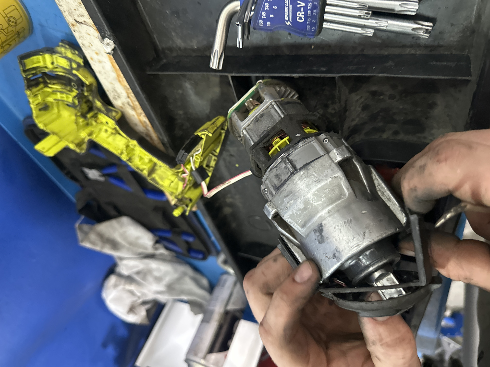The image shows a close-up of a person's hands holding a disassembled electric motor. The motor appears to be part of a power tool, possibly a drill, given the yellow plastic casing visible in the background. The motor itself is made of metal and has wires and internal components exposed. Several Torx bits are visible to the right of the motor, suggesting a repair or maintenance process is underway. The environment appears to be a workshop or garage, with tools and other parts scattered around. There are no visible indications of the location within Blagoveshchensk, Russia.