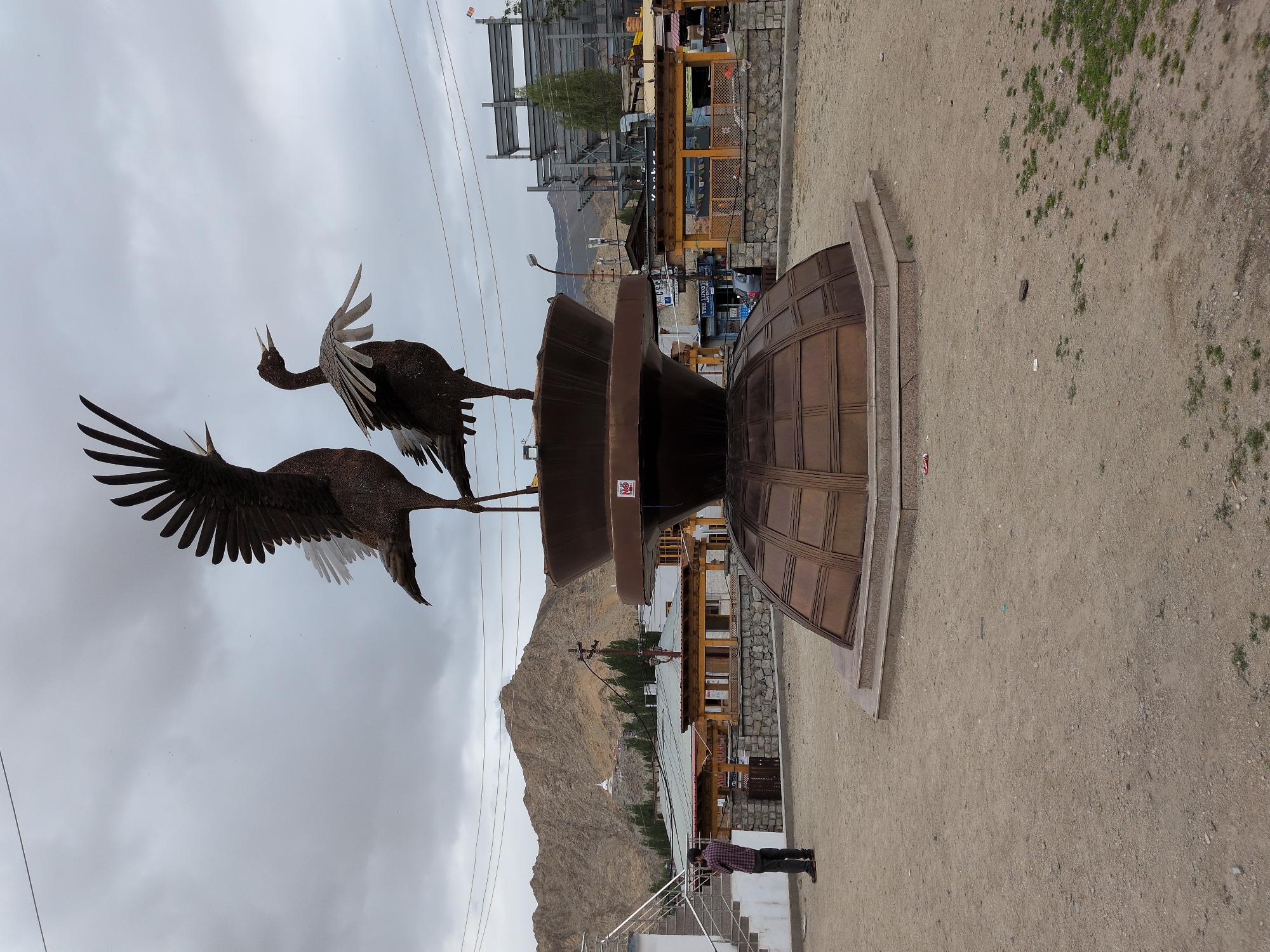 An outdoor, full-frame, eye-level, medium shot shows a large metal sculpture of two birds in a public square in Leh, India. The sculpture depicts two large birds with their wings spread, appearing to be in flight or calling out. They are positioned on top of a tiered, dark bronze pedestal that has a rounded, dome-like base and a wider, bowl-like upper section. The pedestal is situated on a circular stone base. The ground of the square is made of dirt and gravel.
In the background, to the left of the sculpture, a man in a checkered shirt and dark pants is standing and looking at his phone. Behind him, a white building with a peaked roof and a staircase leading up to it is visible, along with a few other low-rise buildings with wooden roofs and stone walls. Further in the background, a large, barren, brown mountain dominates the skyline, with a white stupa or temple structure visible near its peak. Trees with green foliage are scattered across the landscape.
To the right of the sculpture, more low-rise buildings with roofs can be seen, along with some construction in progress, featuring a metal framework structure. Power lines and utility poles are visible throughout the scene, and the sky is overcast with clouds. The overall impression is a blend of public art, urban infrastructure, and natural landscape characteristic of a town in a mountainous region.