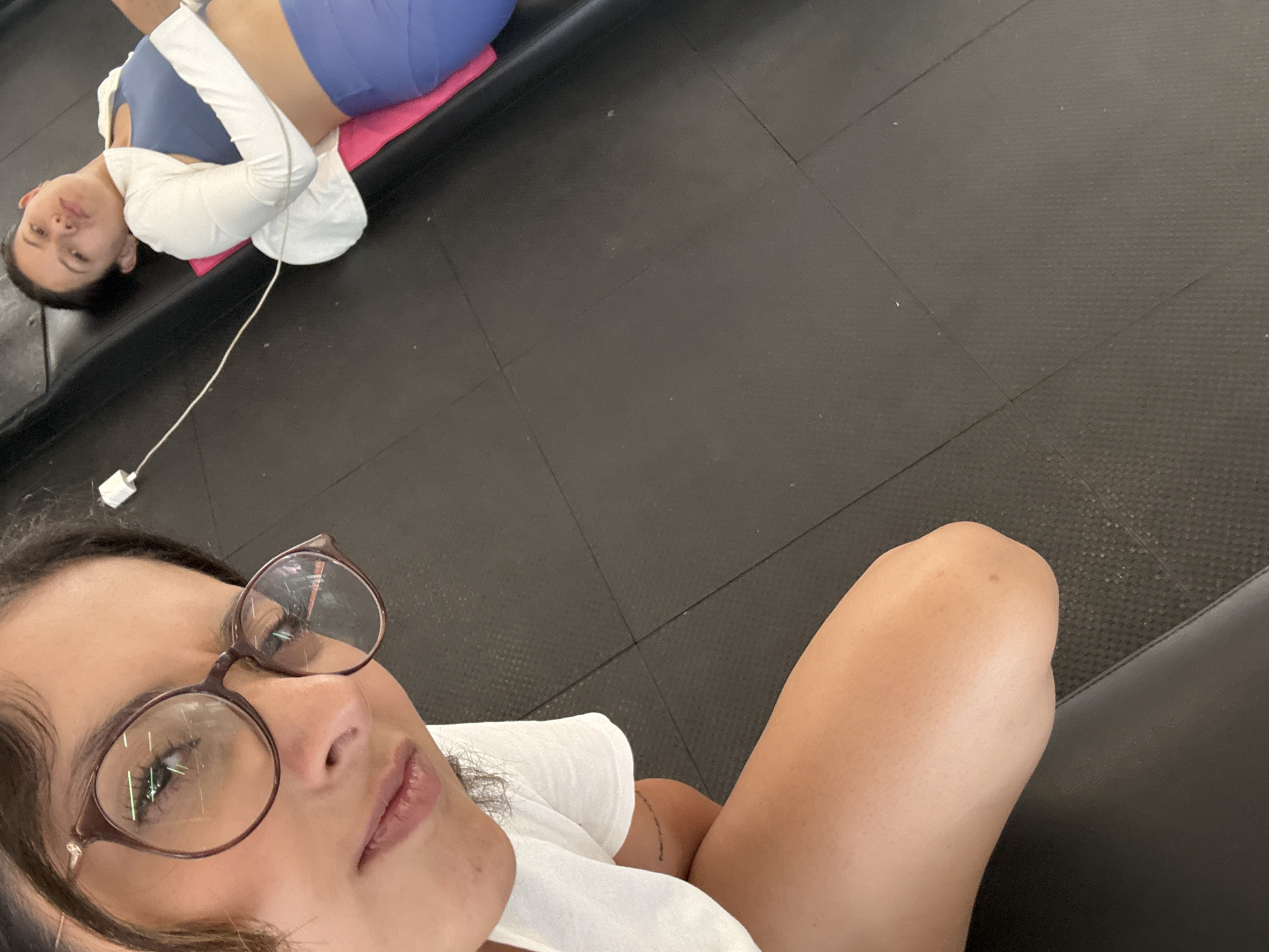 The image depicts two women inside a fitness center in San Andrés Cholula, Mexico. The floor is covered in dark, rubberized tiles. One woman is lying on her back on a pink mat, wearing blue shorts, a blue sports bra, and a white long-sleeved shirt. An earbud cable is visible, connected to a device out of frame. The second woman is closer to the camera, facing the viewer. She is wearing round glasses and a white shirt. Part of her arm and shoulder are visible.