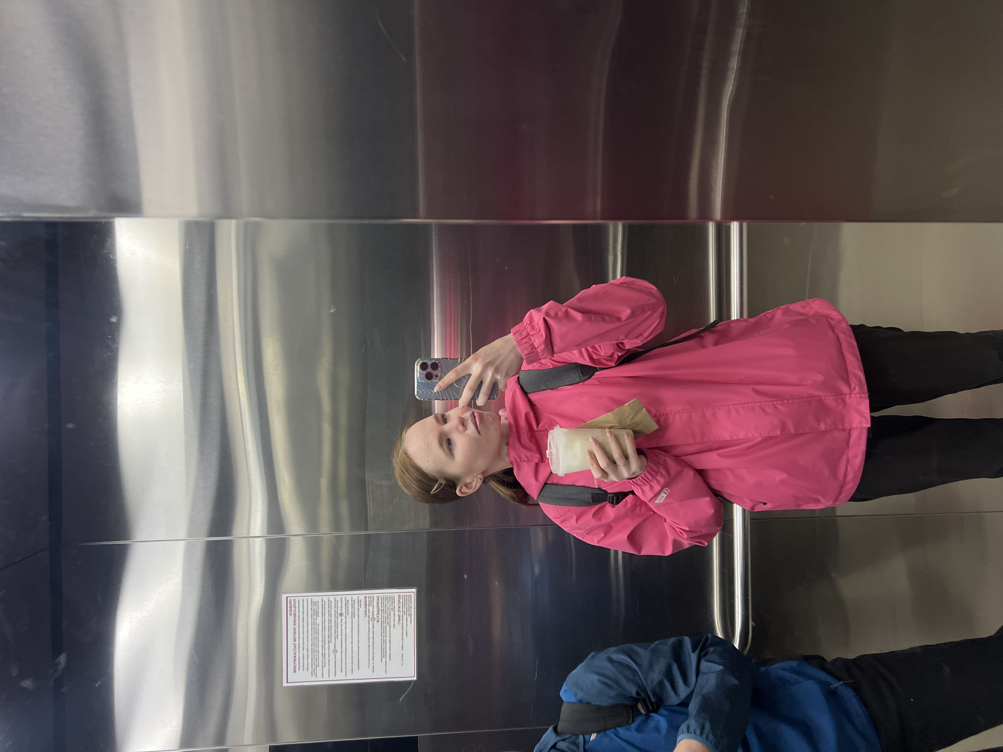 A photograph taken indoors, rotated 90 degrees clockwise, features a mirrored scene of people in what appears to be an elevator. The setting is likely in Perm, Russia, given the Russian text on a sign visible on the elevator wall.
One person, a young adult, is taking a selfie with a smartphone in a mirrored elevator. They are wearing a pink jacket and black pants, and holding a container. There is another individual visible below the first person, wearing a blue jacket and black pants. The elevator's interior consists of metallic surfaces, with vertical lines running along the walls. A sign with Russian text is affixed to the elevator wall.