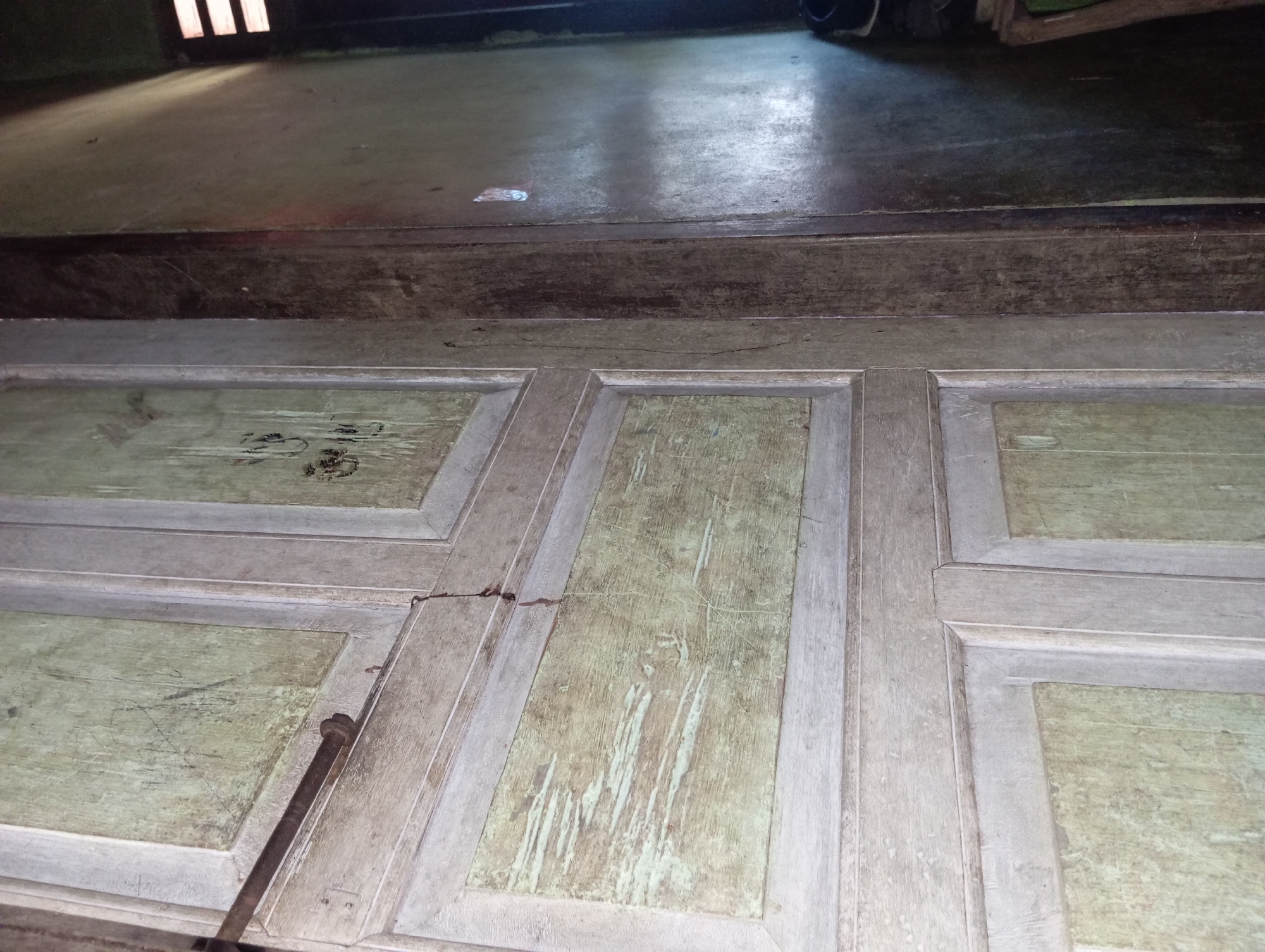The image shows a close-up of an old wooden door with a distressed green and grey painted finish. The door appears to be segmented into panels with raised trim. There is a dark metal rod or pipe resting horizontally across the upper left portion of the door, secured by a bolt or nail. The wood shows signs of wear, with scratches and peeling paint. To the right of the door, a dark, textured wall is visible, possibly made of wood or a similar material. The lighting in the scene suggests an indoor environment, possibly a dimly lit room. There are no people visible in the image. The overall impression is one of age and neglect. The location is identified as Rejosari, Indonesia.
