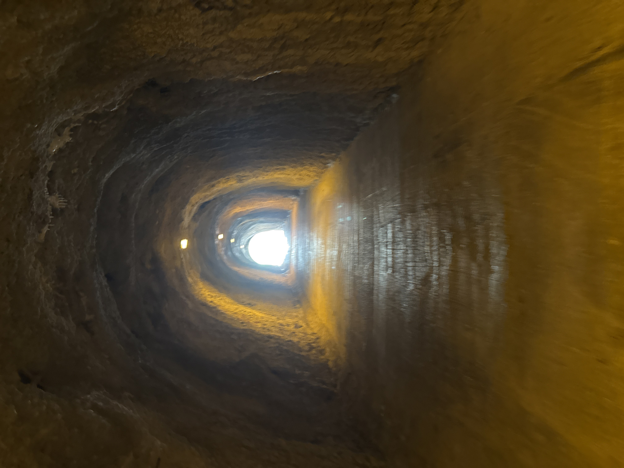 The image depicts the interior of a rough-hewn tunnel, likely carved from rock. The tunnel walls are uneven and textured, with a distinctly organic, almost cave-like appearance. The lighting is dim, originating from a bright, diffused light source at the far end of the tunnel, creating a strong sense of depth and perspective. Several small, yellowish lights are visible along the walls closer to the viewer, providing ambient illumination. The overall color palette is dominated by earthy browns, grays, and warm yellows, enhanced by the glow emanating from the tunnel's exit. The tunnel appears to be an enclosed, man-made or natural passage, leading towards an opening. The texture of the walls suggests it could be a historical site, perhaps a fortification, an ancient passage, or a natural geological formation adapted for use. There is no visible text in the image, nor are there any people or specific objects that clearly identify the purpose or exact nature of the tunnel, other than its general form as an underground passageway. The mood is somewhat mysterious and atmospheric, with the bright light at the end drawing the viewer's eye and suggesting a transition to an external space.