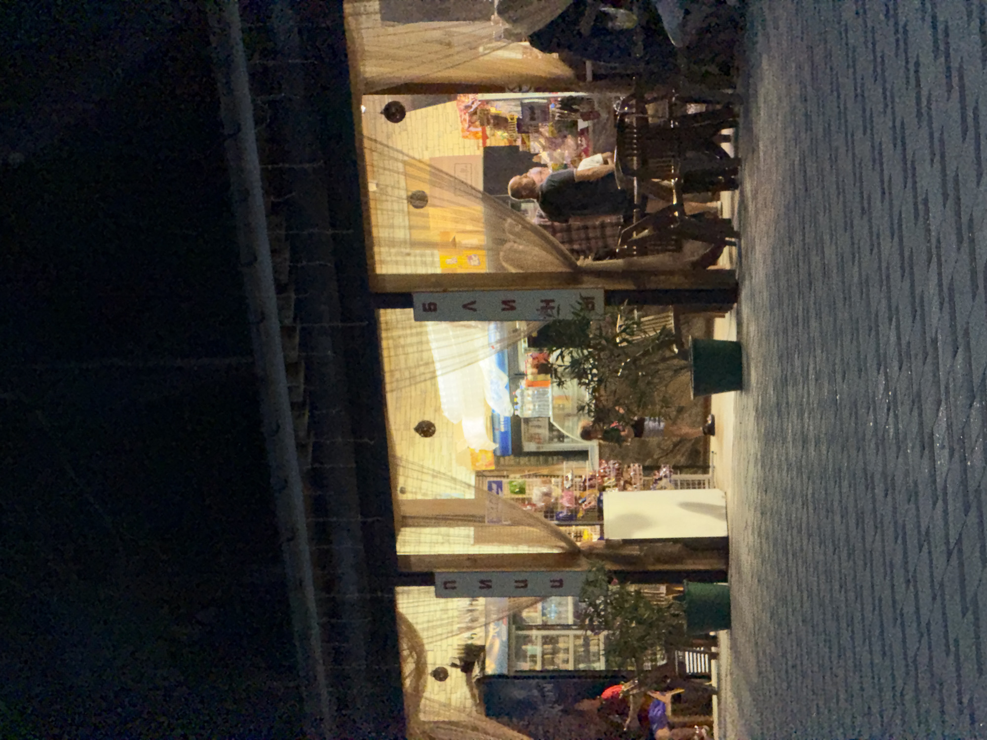 The image shows an outdoor scene at night. In the foreground, there is a dark, textured surface, possibly a wall or pavement. To the right, a paved area with a distinctive diamond pattern suggests a public space like a sidewalk or plaza.
The main focus is a dimly lit shop or cafe, viewed through a translucent curtain or screen. Inside, shelves are stocked with various items, suggesting it might be a small store or a market stall. A few people are visible. One man, seated at a table, appears to be looking towards the camera or the entrance. He is wearing a dark shirt and is bald. Further inside, other figures are blurred and indistinct.
A sign with Cyrillic text is partially visible hanging from the ceiling of the shop. The text on the sign appears to be "СУШИ" which means "sushi". Another sign, with characters that are difficult to decipher, is also present.
The lighting is warm and artificial, coming from within the establishment and some overhead lights. The overall atmosphere is relaxed, possibly indicating a casual dining or shopping experience. The time of day is clearly night, and the weather appears to be clear. The scene evokes a sense of local commerce and evening activity. The visual context of the sign and the general ambiance suggest a setting in Partenit, Ukraine.