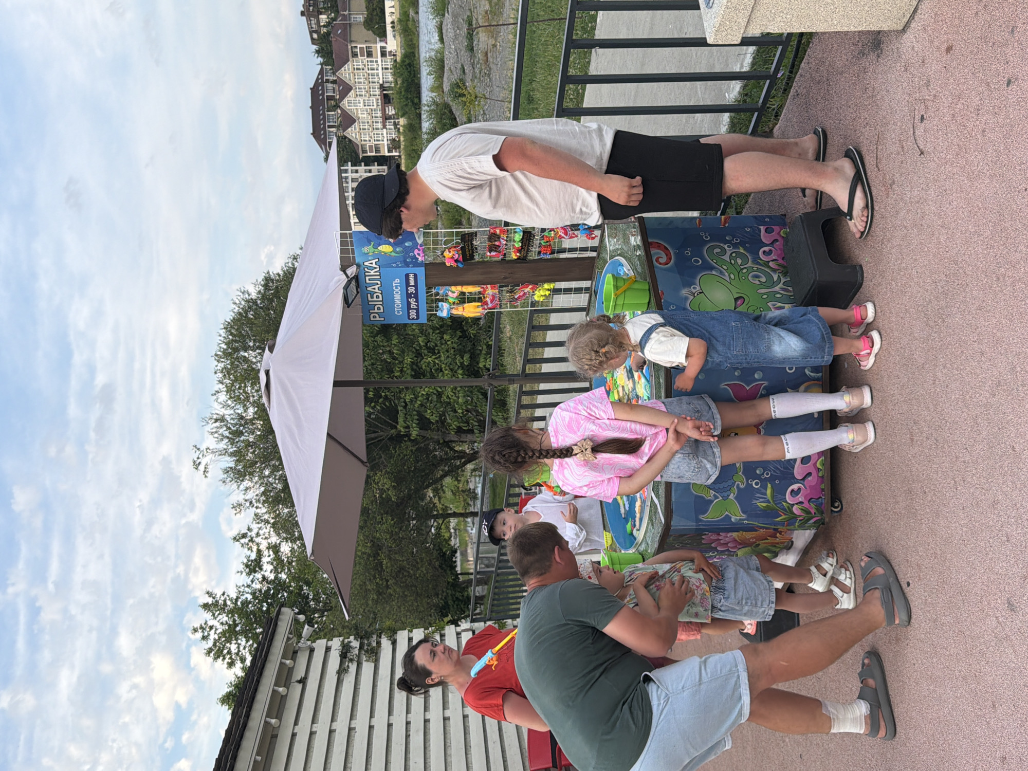 This image depicts a sunny outdoor scene with several people, including adults and children, gathered around a carnival game stall. The stall, which appears to be a fishing game, is decorated with aquatic-themed graphics. A sign on the stall reads "РЫБАЛКА" (Fishing) and indicates a cost of "300 руб - 30 мин" (300 rubles - 30 minutes). Various small toys or prizes are displayed on racks next to the stall.
In the foreground, a man in a green t-shirt and light blue shorts is bending down, interacting with a young child wearing a floral top and denim overalls. To the right of them, a young girl with braided hair in a pink t-shirt and denim shorts stands with her hands behind her back, facing the game. Another child in a white shirt and a baseball cap is visible behind them. A woman in a red t-shirt is standing to the left, holding a yellow and blue toy. A man in a white t-shirt and black shorts, wearing a black cap, is observing the game.
The environment suggests a recreational area, possibly a park or waterfront. In the background, lush green trees and residential buildings can be seen, consistent with the landscape of Sochi, Russia, a coastal city known for its resort areas. The sky is partly cloudy, indicating a pleasant day. A metal railing runs along the edge of the area, and a white building with horizontal siding is present on the left. The ground is paved with a reddish-pink material.