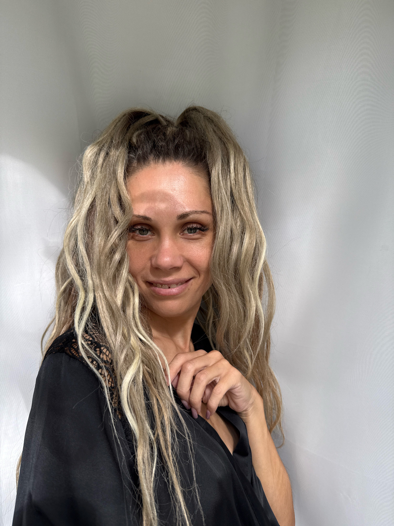 A medium shot shows a woman with blonde, wavy hair pulled into a high ponytail. She is wearing a black robe with lace accents. Her right hand is near her neck, and she is looking at the camera with a slight smile. The background is a plain white curtain. The image appears to be taken indoors, with soft lighting. There is no information to suggest the location is in Rusanovo, Russia.