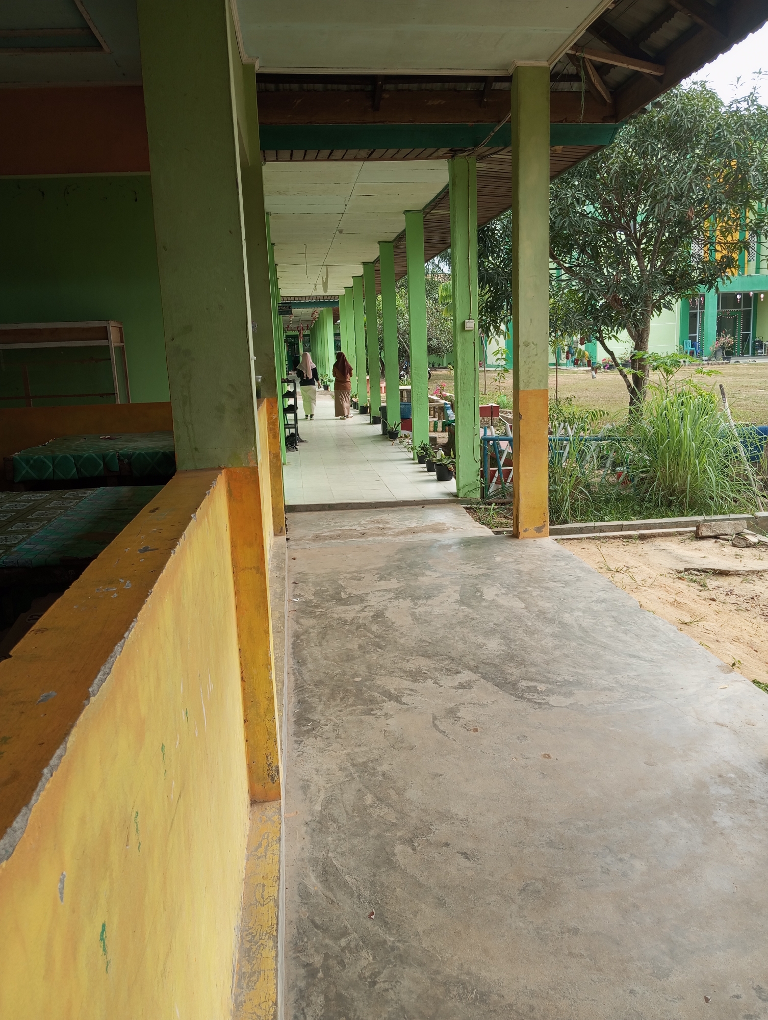 The image captures a view down a covered walkway with a concrete floor. The walkway is bordered on the left by a yellow-painted wall and on the right by green-painted pillars supporting a wooden roof. In the background, two individuals are walking away from the camera, one wearing a white hijab and the other a brown headscarf. Beyond the walkway, a grassy area is visible with trees and a building with green and yellow accents. The scene appears to be an outdoor corridor at a school or public institution in Pematang Reba, Indonesia, likely during daylight hours given the ambient light. The walkway is empty except for the two individuals in the distance. The environment is moderately tidy, with some vegetation and sandy ground visible to the right.