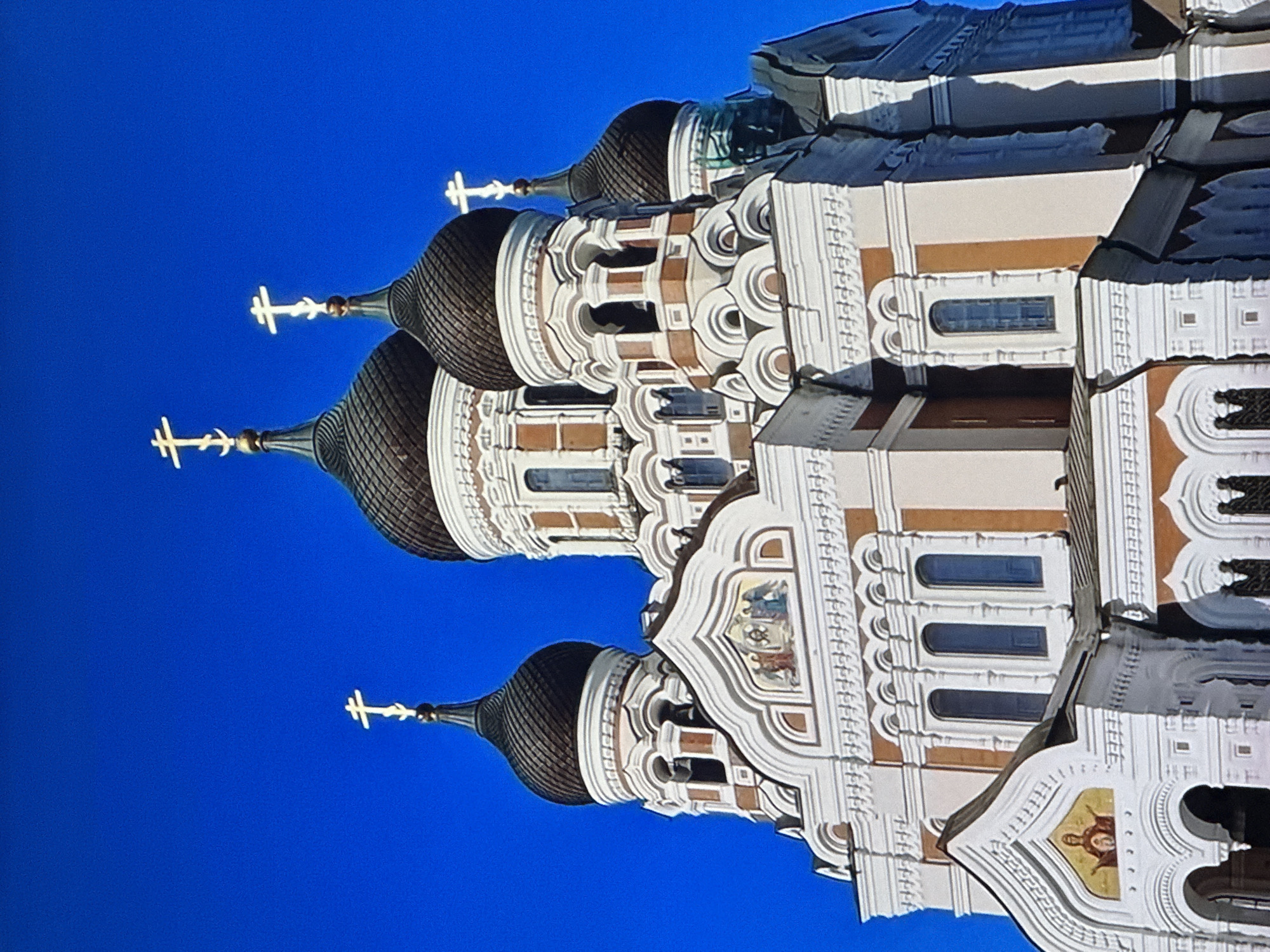 This is a low-angle, close-up, eye-level, outdoor, daylight photograph of the Alexander Nevsky Cathedral in Tallinn, Estonia. The cathedral is a Russian Orthodox cathedral with multiple onion domes topped with golden crosses. The building is predominantly white with terracotta accents and intricate architectural details. The sky is a clear, vibrant blue. There are no people visible in the image. The composition focuses on the upper sections of the cathedral, highlighting the domes, crosses, and facades.