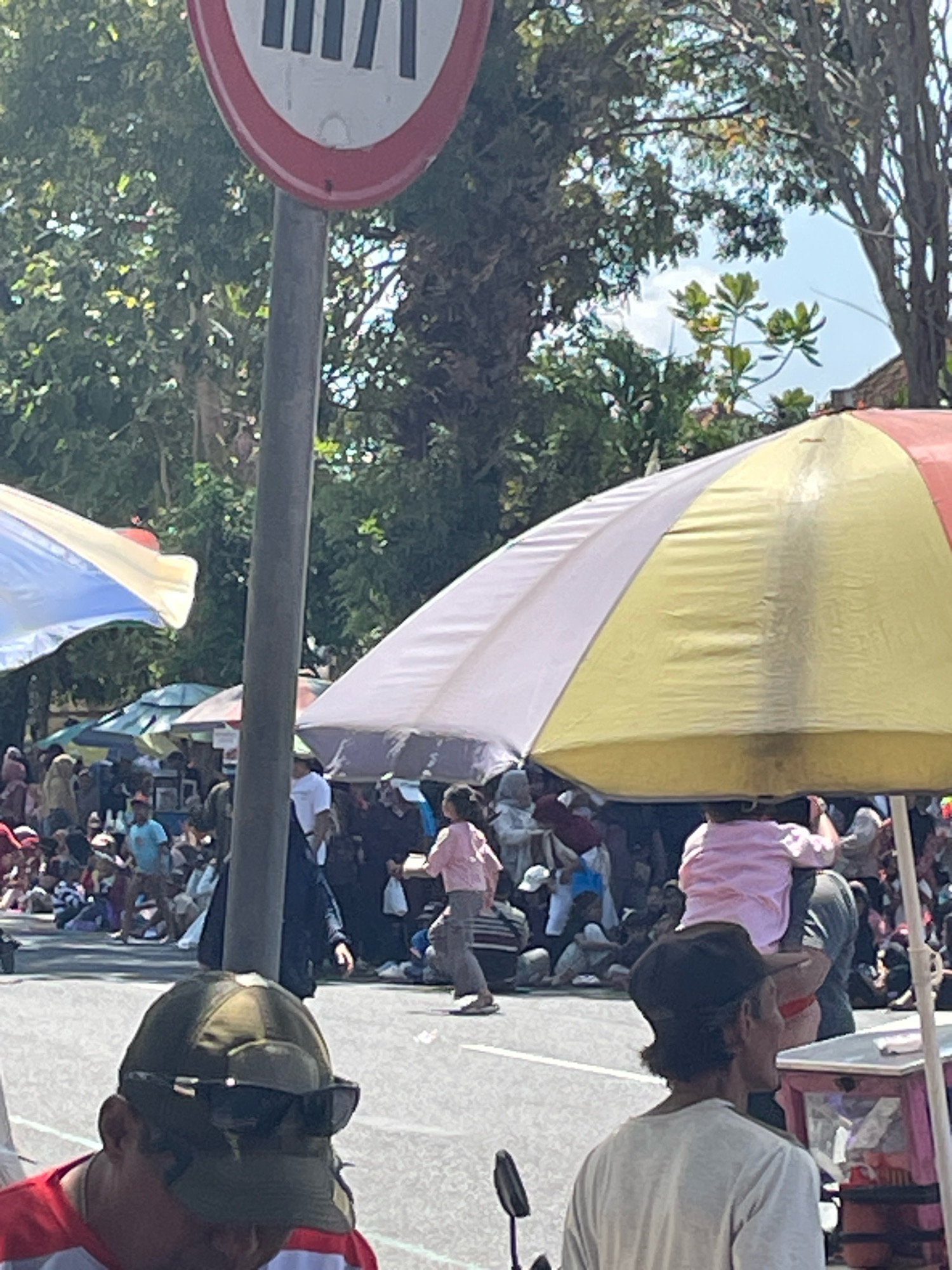This image depicts a street scene in Purworejo, Indonesia, likely during a public event or festival. In the foreground, a man wearing a green cap and sunglasses is partially visible, looking downwards. Behind him, another man in a white t-shirt faces away from the camera. A large, colorful umbrella, segmented in white, yellow, and red, dominates the right side of the frame, casting shade.
Further back, a crowd of people are gathered, many sitting or standing, suggesting a parade, market, or gathering. A young child in a pink shirt is being carried on someone's shoulders amidst the crowd. Various stalls and smaller umbrellas are visible among the attendees, indicating a bustling outdoor environment. Trees and the sky are visible in the background, suggesting it is daytime. A traffic sign with red and white markings is positioned on the left side of the image, affixed to a pole. The ground appears to be asphalt or a similar paved surface. The overall atmosphere suggests a lively public event in an urban or semi-urban setting.