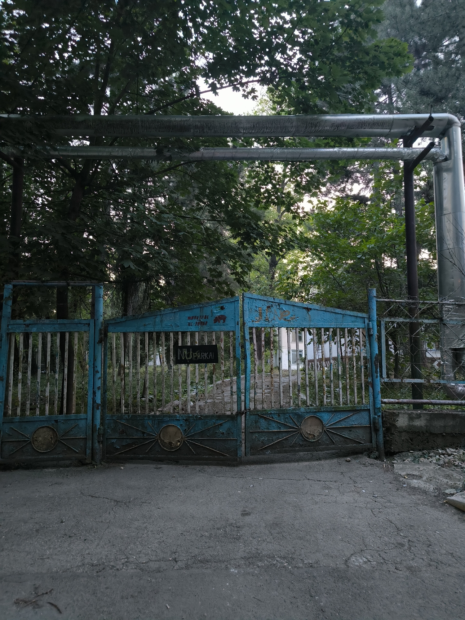 The image depicts an old, turquoise-painted metal gate in Chișinău, Moldova. The gate is slightly ajar and features a faded inscription on the left panel that appears to be a Cyrillic alphabet word, possibly "Парковка" (Parkovka), meaning "parking." A smaller black sign with white lettering, reading "NU PARKA," is attached to the right gate panel. Behind the gate, overgrown vegetation is visible, with some hints of buildings in the distance. Two large metal pipes run horizontally across the upper portion of the frame, supported by posts and stretching over the gate and trees. The ground in front of the gate is paved with asphalt. The overall scene suggests a somewhat neglected or abandoned area within the city.