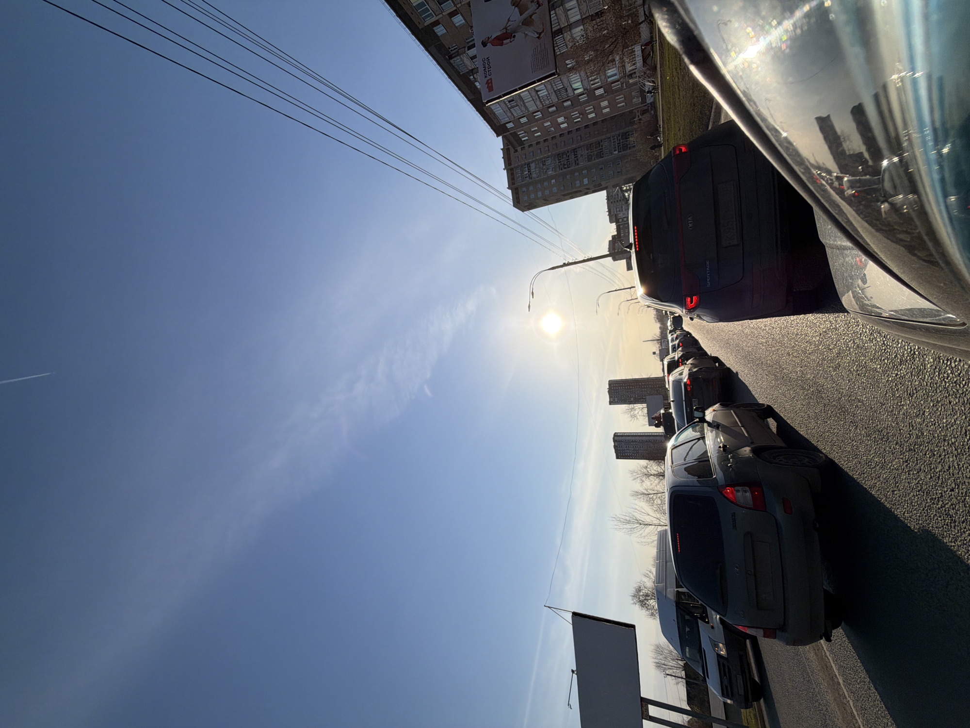 The image shows a traffic jam on a sunny day in a city. Cars are lined up on the asphalt road, reflecting sunlight. Buildings are visible in the background, including a high-rise structure with advertisements. Overhead power lines run across the top of the image. A blank billboard stands along the roadside. There are apartment buildings in the upper part of the image as well. Based on the architecture and the presence of a Lada car, the location is Izhevsk, Russia.