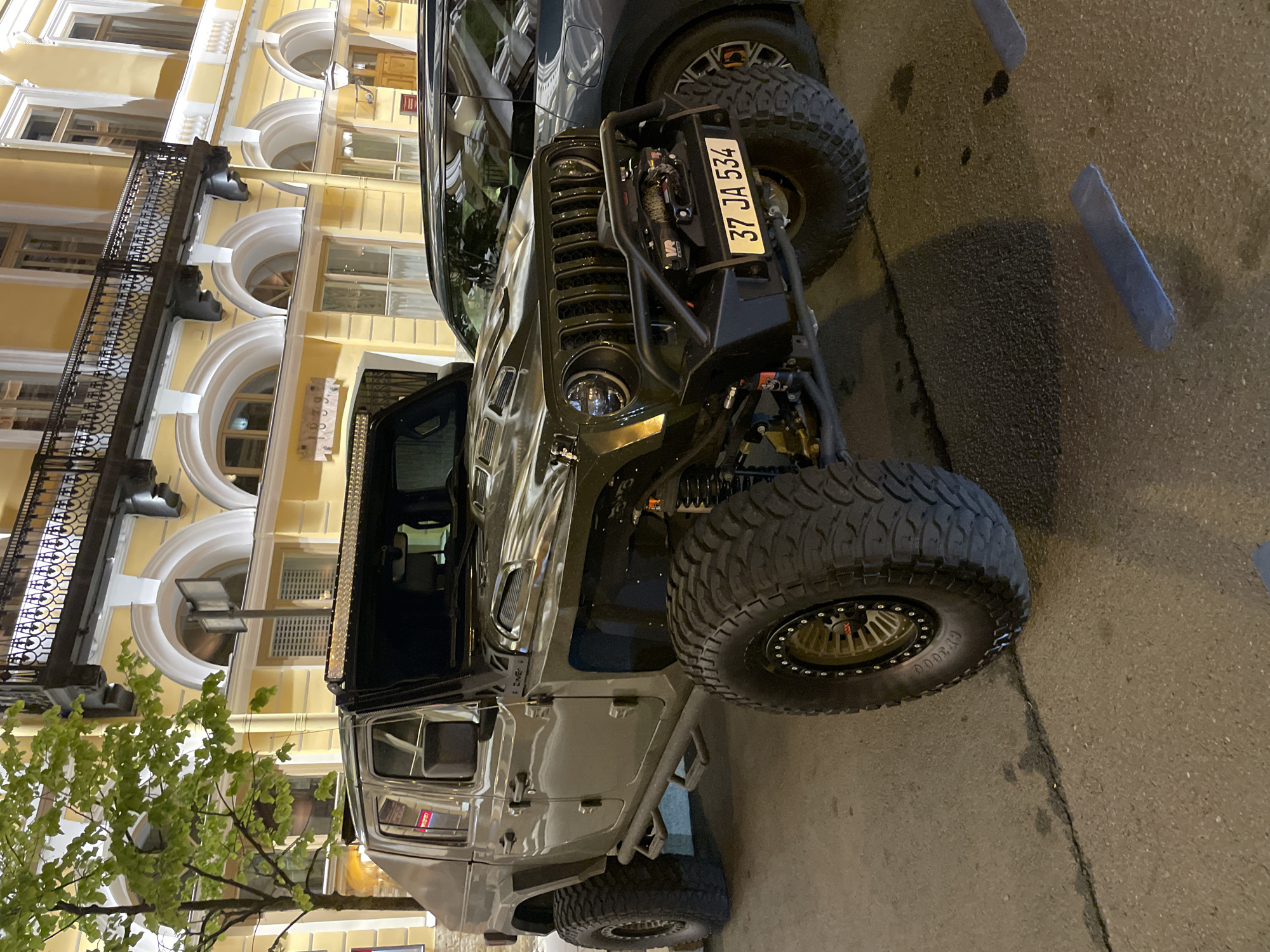 The image shows a dark gray Jeep Wrangler, heavily modified for off-road use, parked on a street in Saint Petersburg, Russia. The vehicle has oversized tires, a custom front bumper with a winch, and additional protective bars. The license plate reads "37 JA 534". The vehicle is parked near a yellow building with arched windows and decorative iron balconies, characteristic of Saint Petersburg architecture. A tree with green leaves is visible on the left side of the frame. The lighting suggests it is evening. There are parking blocks on the right side of the street.