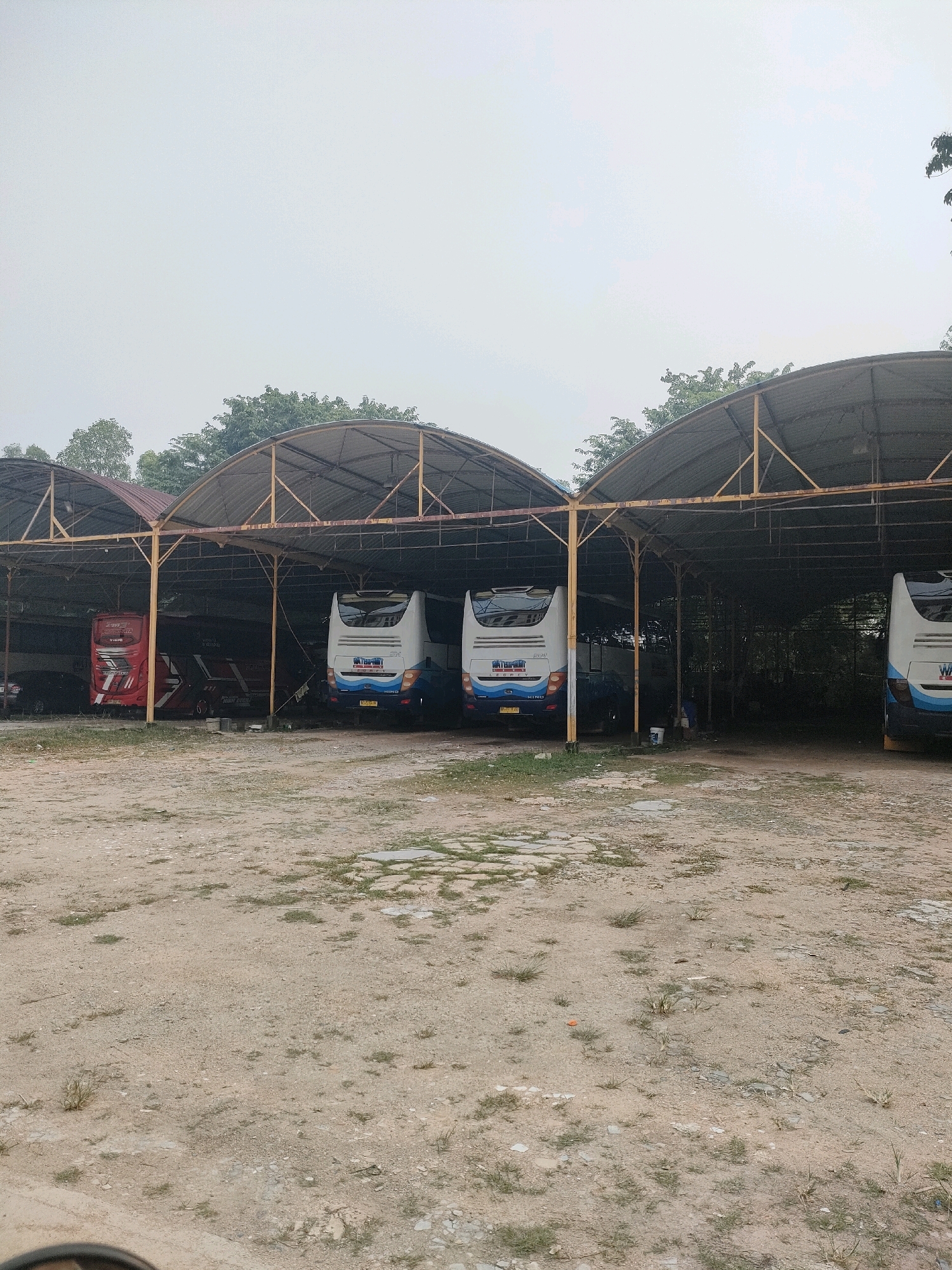 A view of a bus depot in Batam, Indonesia shows several buses parked under a weathered open-sided metal shelter. The shelter has a curved, corrugated metal roof supported by yellow metal poles and beams. The ground is unpaved, consisting of dirt and gravel with patches of sparse grass and some scattered flat stones. In the background, green trees are visible beyond the shelter. The sky is overcast and a pale gray.
The buses are primarily white with blue and red accents. Two white buses with blue stripes are parked side-by-side under the main section of the shelter, their rear ends facing the camera. One of these buses displays a logo with blue and white lettering. To the left, a red bus with white and black graphics is parked. There are other vehicles and objects visible under the shelter in the background, but they are not clearly identifiable due to shadow and distance. One of the white buses has a license plate that is partially obscured. A red dispenser-like object is visible to the far left, near the red bus. There are some buckets or containers on the ground in the middle of the depot. The scene appears to be of a functional, somewhat rustic, bus parking or maintenance area.