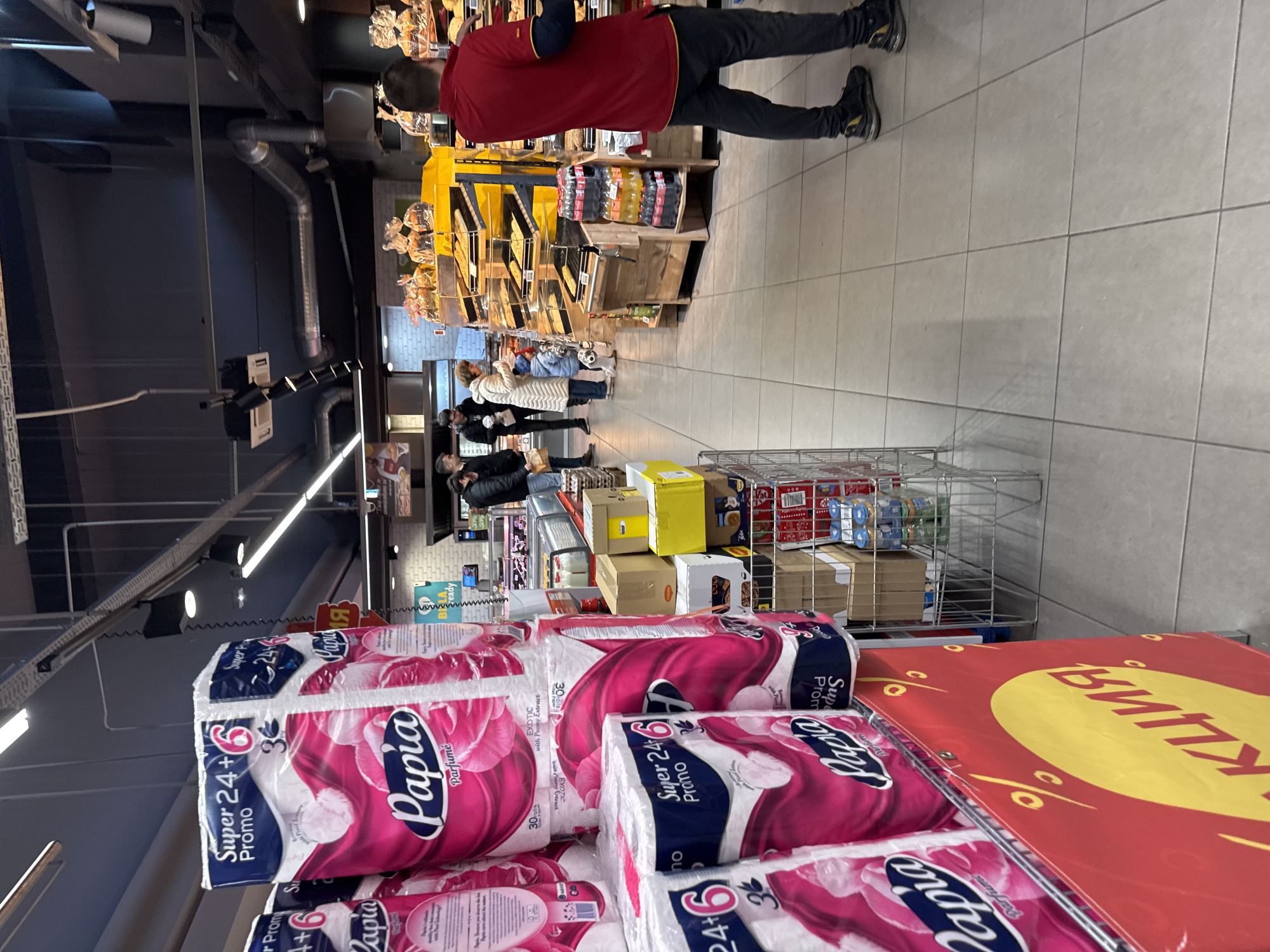 Here's a matter-of-fact description of the image:
**Scene:** The image depicts the inside of a grocery store, likely in Sofia, Bulgaria, due to the presence of Cyrillic script on a promotional sign.
**Foreground:** There is a shopping cart filled with "Papia" brand toilet paper. The packaging indicates a "Super 24+6 Promo." A red and yellow promotional sign is near the toilet paper.
**Midground:** A metal shopping cart further down the aisle is filled with cardboard boxes and some packaged food items. Several people are visible in the aisle beyond the carts, seemingly browsing and shopping.
**Background:** The aisle extends into the distance with a bakery display on the left, staffed by a person in a red shirt, and shelving stocked with products. The store has overhead lighting and exposed ductwork.
**Overall Impression:** The scene shows an average shopping environment with shoppers, groceries, and promotional displays.