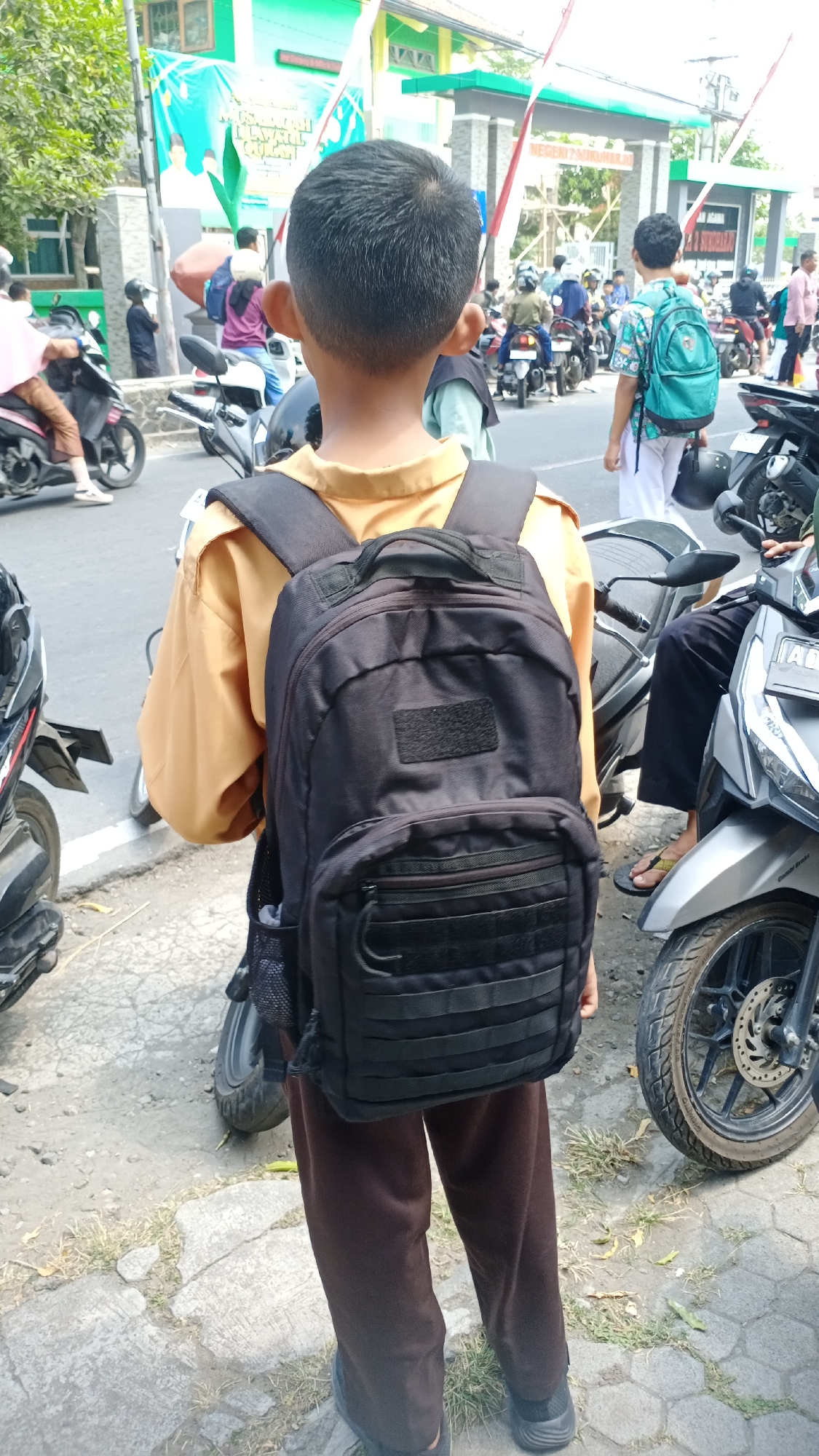 A young male wearing a tan uniform shirt and dark brown pants stands facing away from the camera, carrying a black backpack. He is on a paved road with a sidewalk to his left. Several motorcycles are parked on the road, and other people are visible in the background, some riding motorcycles. A building with a green roof and a large banner is in the background, with what appears to be a school entrance. The scene is consistent with a school setting in a tropical climate, likely during the day. The location is specified as Joho, Indonesia.