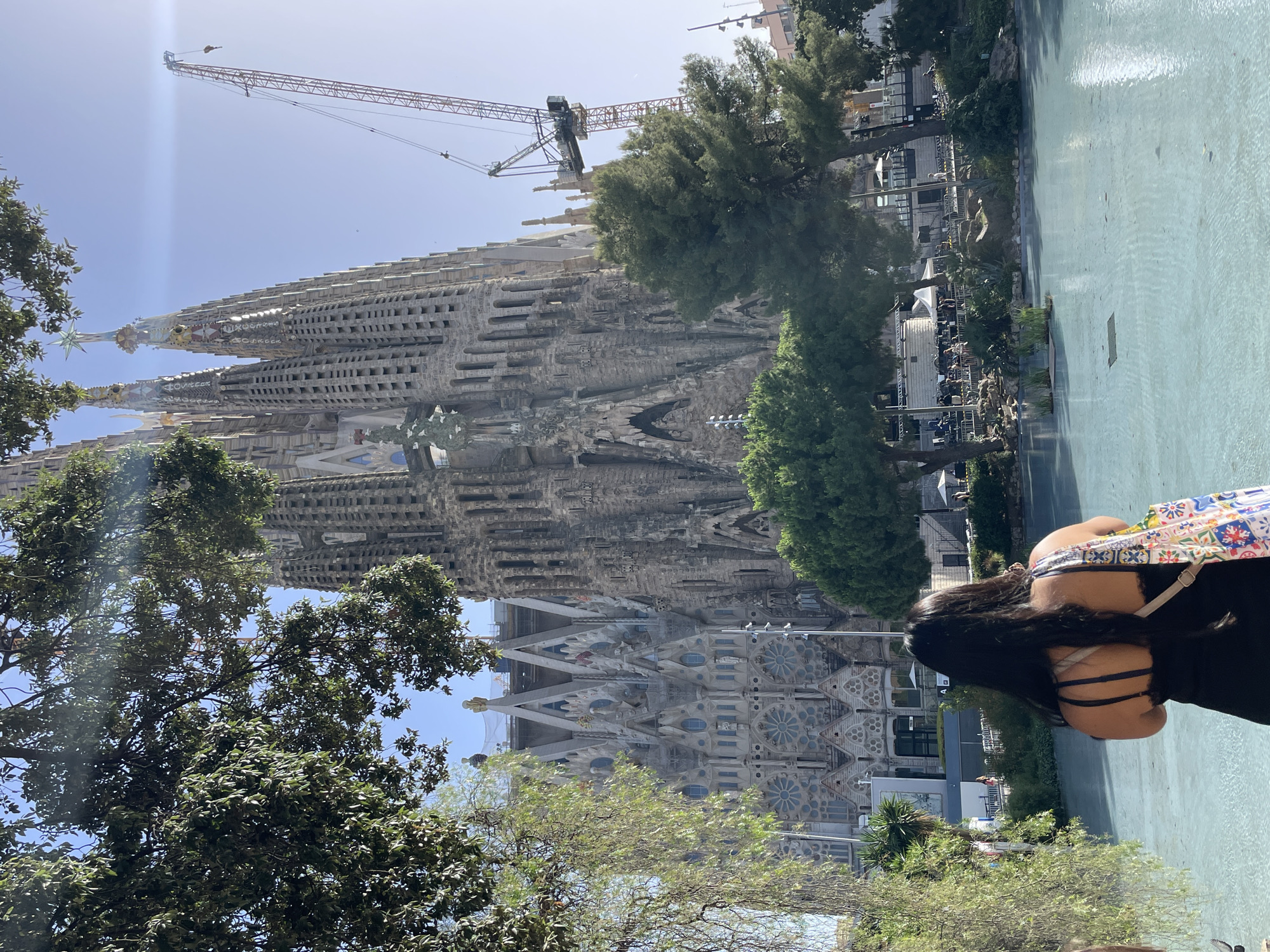 The image depicts the Sagrada Familia in Barcelona, Spain. In the foreground, a woman with long dark hair faces away from the camera, wearing a black top and carrying a colorful tote bag. She stands near a pale blue body of water. Behind her, the Sagrada Familia's unfinished facade is visible, characterized by its intricate stone carvings and tall spires. A large orange construction crane is positioned to the right of the basilica, indicating ongoing construction. Lush green trees and foliage surround the structure and the water feature, with a bright sky overhead. The scene appears to be a daytime exterior shot.
