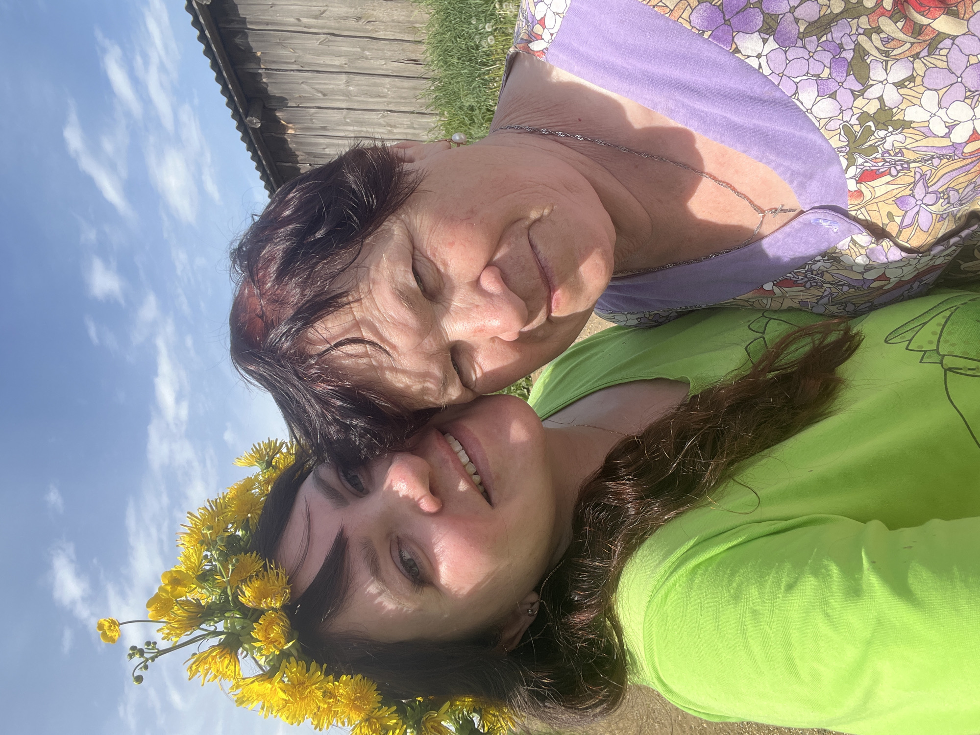 Two women are lying on the ground outside. One woman, likely older, wears a lavender floral top and has her eyes closed, resting her face close to the younger woman. The younger woman, wearing a bright green shirt and a dandelion wreath in her hair, smiles at the camera. In the background, there is a wooden structure and a blue sky with clouds. Although not verifiable with the imagery, the location is implied to be Panteleevka, Russia.