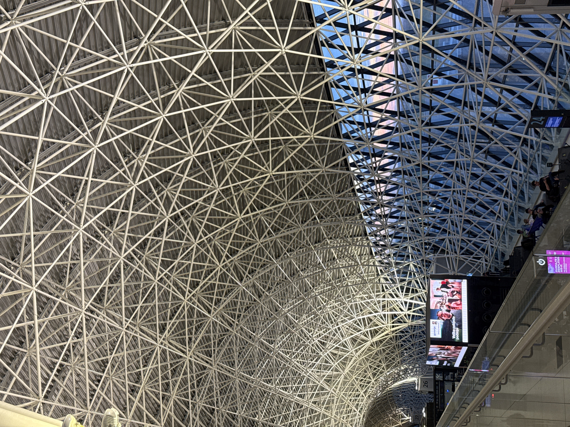 The image shows the interior of Zagreb Airport, located in City of Velika Gorica, Croatia. The ceiling is composed of a complex, interwoven lattice of white metal beams, creating a geometric pattern. Some sections of the ceiling are made of transparent glass, allowing natural light to filter through. In the lower-right quadrant of the image, there are signs with text and numbers, along with a digital advertisement screen. There are a few blurred people visible on an upper level next to the right edge of the frame. The overall tone is modern and architectural.