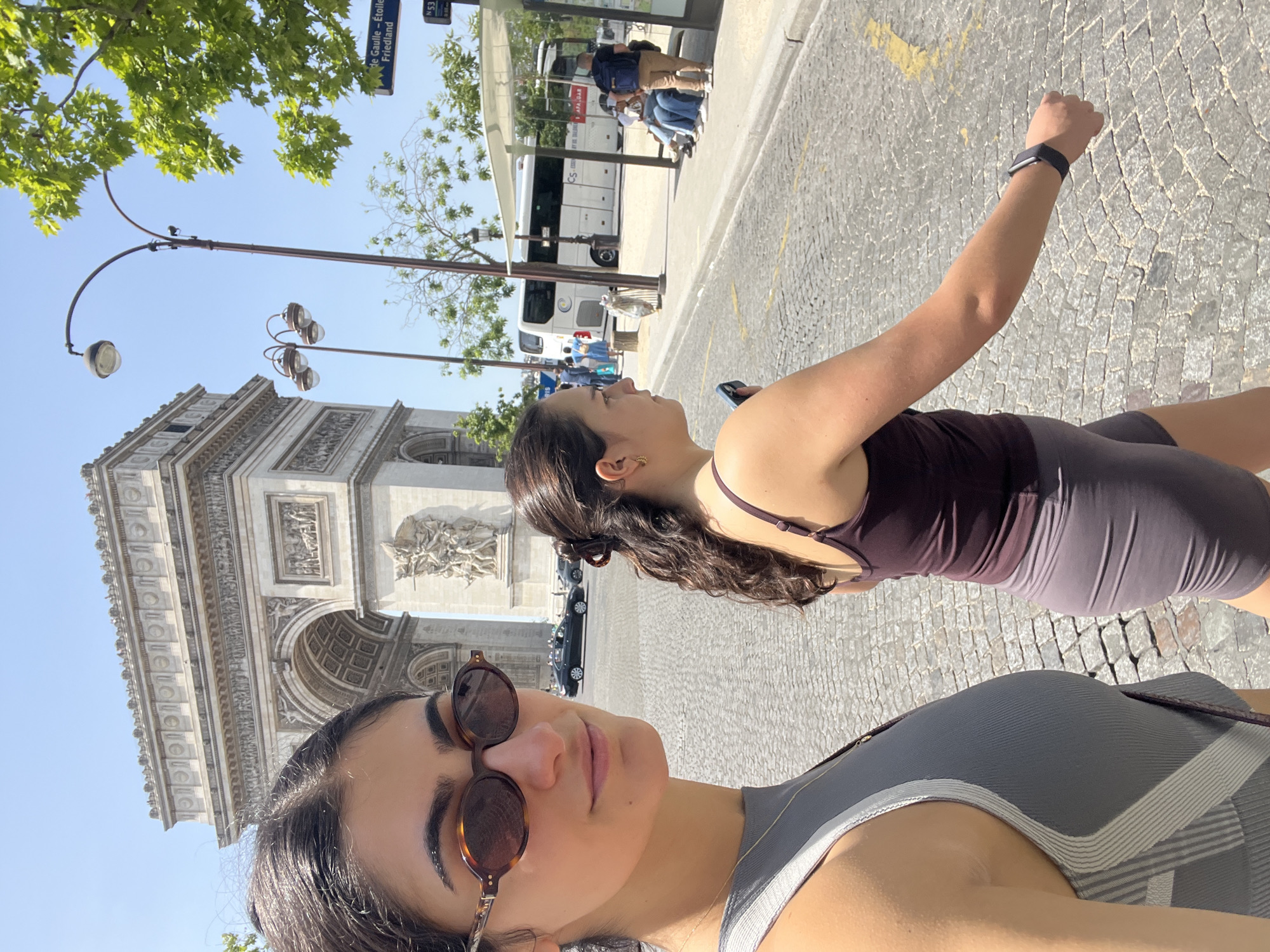 Two women are posing for a selfie in front of the Arc de Triomphe in Paris, France. The woman in the foreground is wearing sunglasses and a grey tank top. The woman behind her is wearing a maroon top and grey athletic shorts, and she has an Apple Watch on her left wrist. A street sign is visible in the top left of the frame, reading "de Gaulle-Etoile Friedland". Behind the women, there is a bus stop where people are waiting. The day is sunny, and the sky is blue.