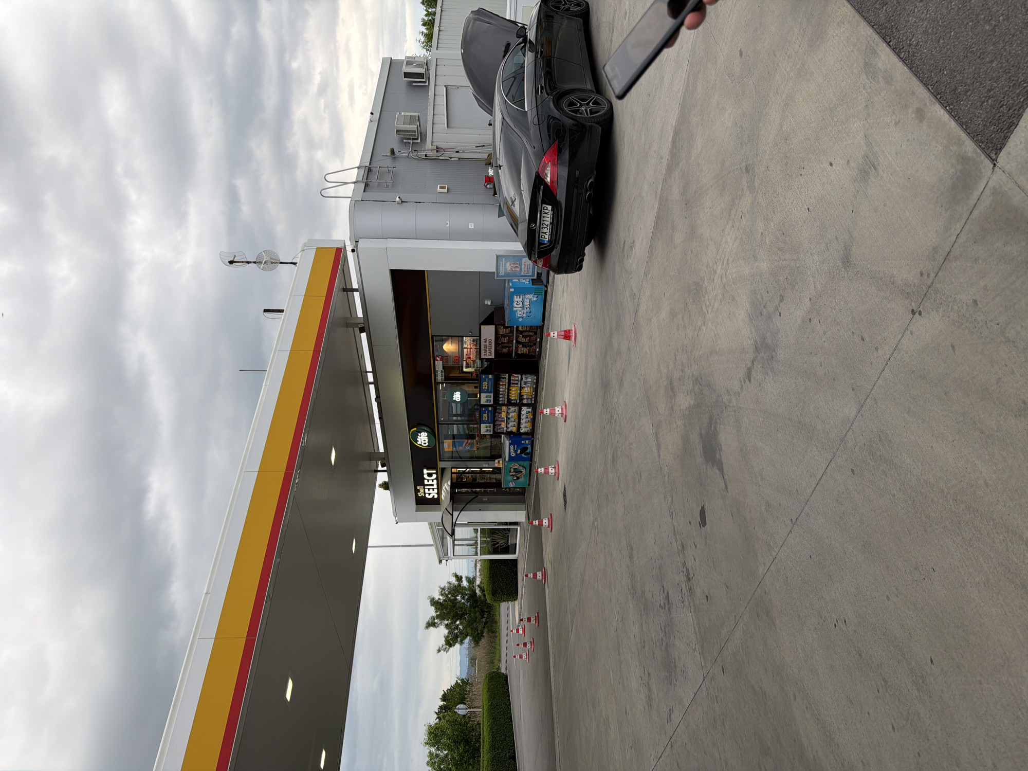 A black Mercedes-Benz with license plate PA3211KP is parked at a Shell gas station convenience store labeled "SELECT" in Dragor, Bulgaria. The hood of the car is open. Orange and white traffic cones delineate a path along the gas station's concrete apron. A person is holding a cell phone in the upper right corner of the image. The sky is overcast.