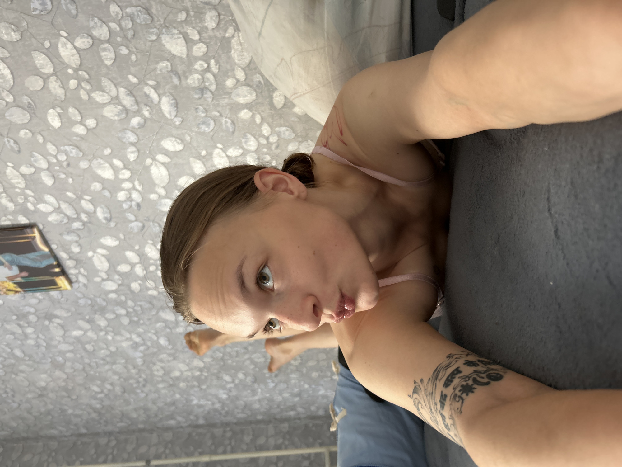 The image shows a young woman lying on her stomach on a bed with a gray textured wallpaper in the background. The woman is looking to her right with her lips pursed. She has a tattoo on her left arm. There is a pillow behind her head and another pillow visible to the right. The lighting suggests an indoor environment, likely a bedroom. The presence of a textured wallpaper and a bed indicates a residential setting. The woman appears to be at home. There are no clear indications of the specific location within the city of Chaikovsky, Russia, beyond it being an indoor residential scene.