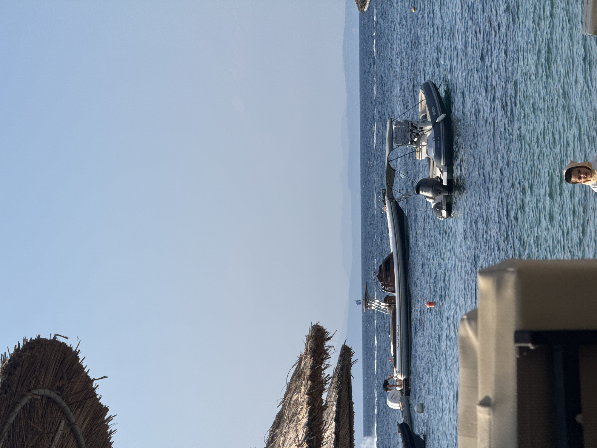 The image depicts a scene at a beach in Sithonia Municipal Unit, Greece. In the foreground, a child wearing a light-colored hooded garment stands in shallow turquoise water. In the middle ground, two inflatable boats are present on the blue sea. The boat on the left appears to be larger and has a person in dark clothing attending to its engine. It displays the brand "CAPELLI." The boat on the right is smaller and has a gray canopy. Further in the background, a coastline with hills is visible, and the sky is a clear, pale blue. Two thatched umbrellas are positioned in the upper left of the frame, suggesting a beach resort setting.