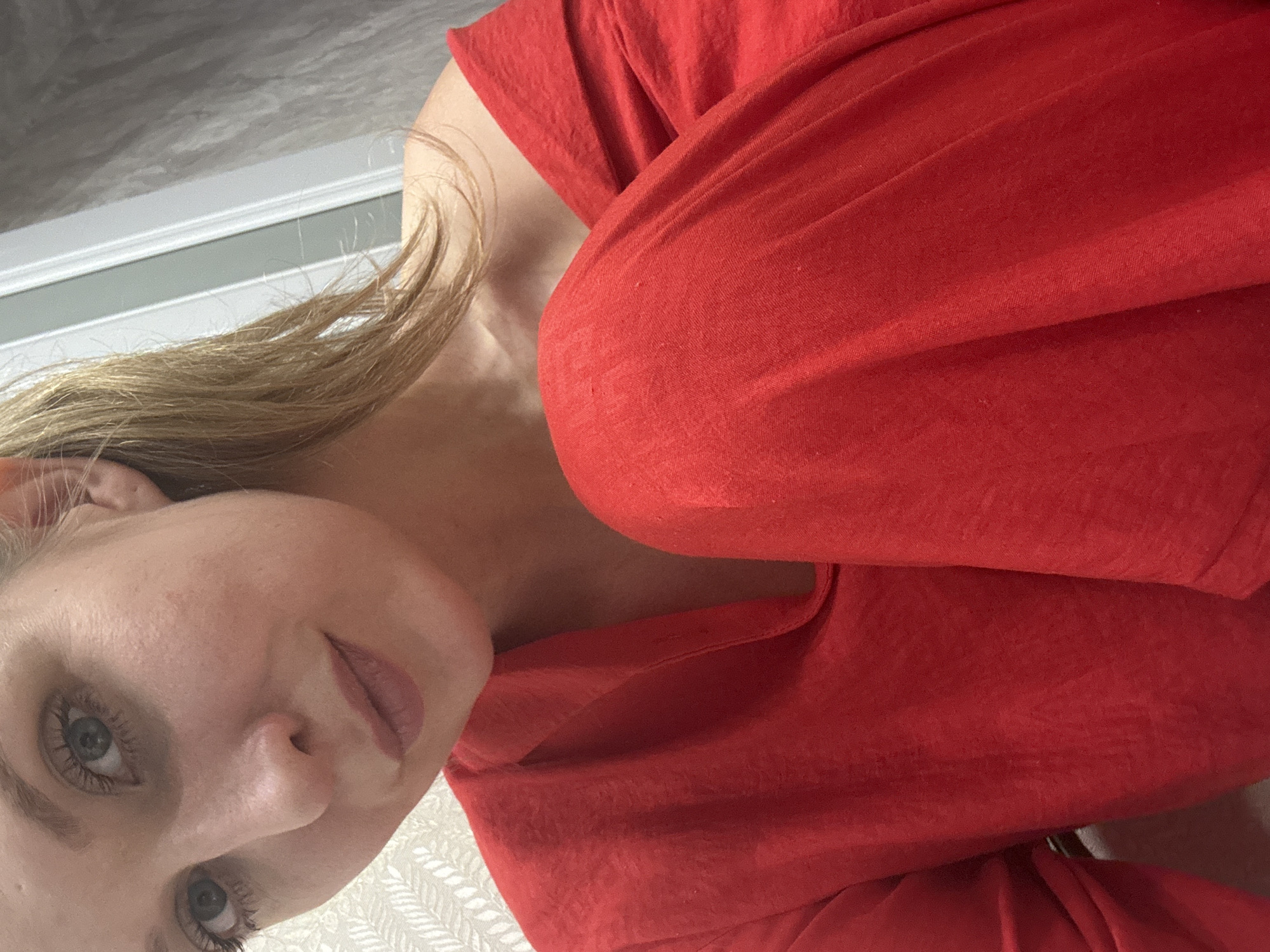 A woman with blonde hair and blue eyes, wearing a red top, is positioned in the frame. The background includes a light gray wall and what appears to be patterned fabric. The scene is not identifiable as being in Izhevsk, Russia, or any other specific location.